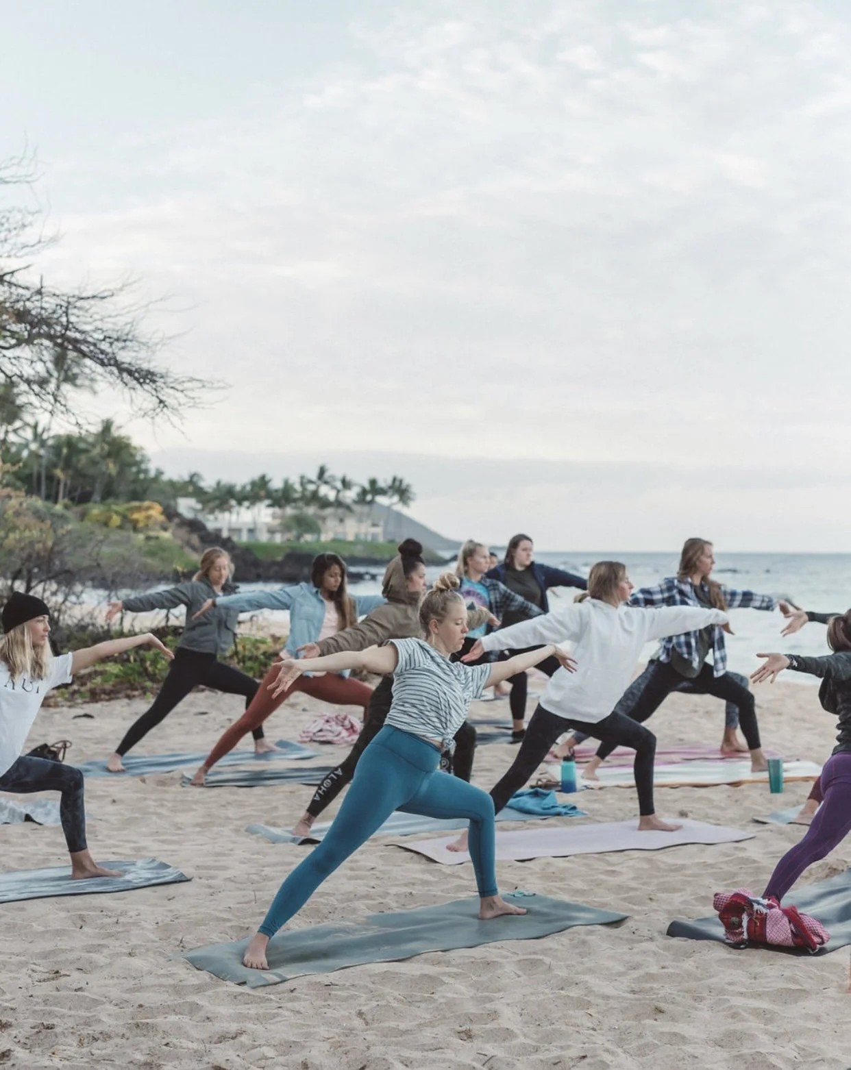 A group of women practicing yoga on a beach, standing on yoga mats with arms extended and one leg bent, near the shoreline with trees and houses in the background.