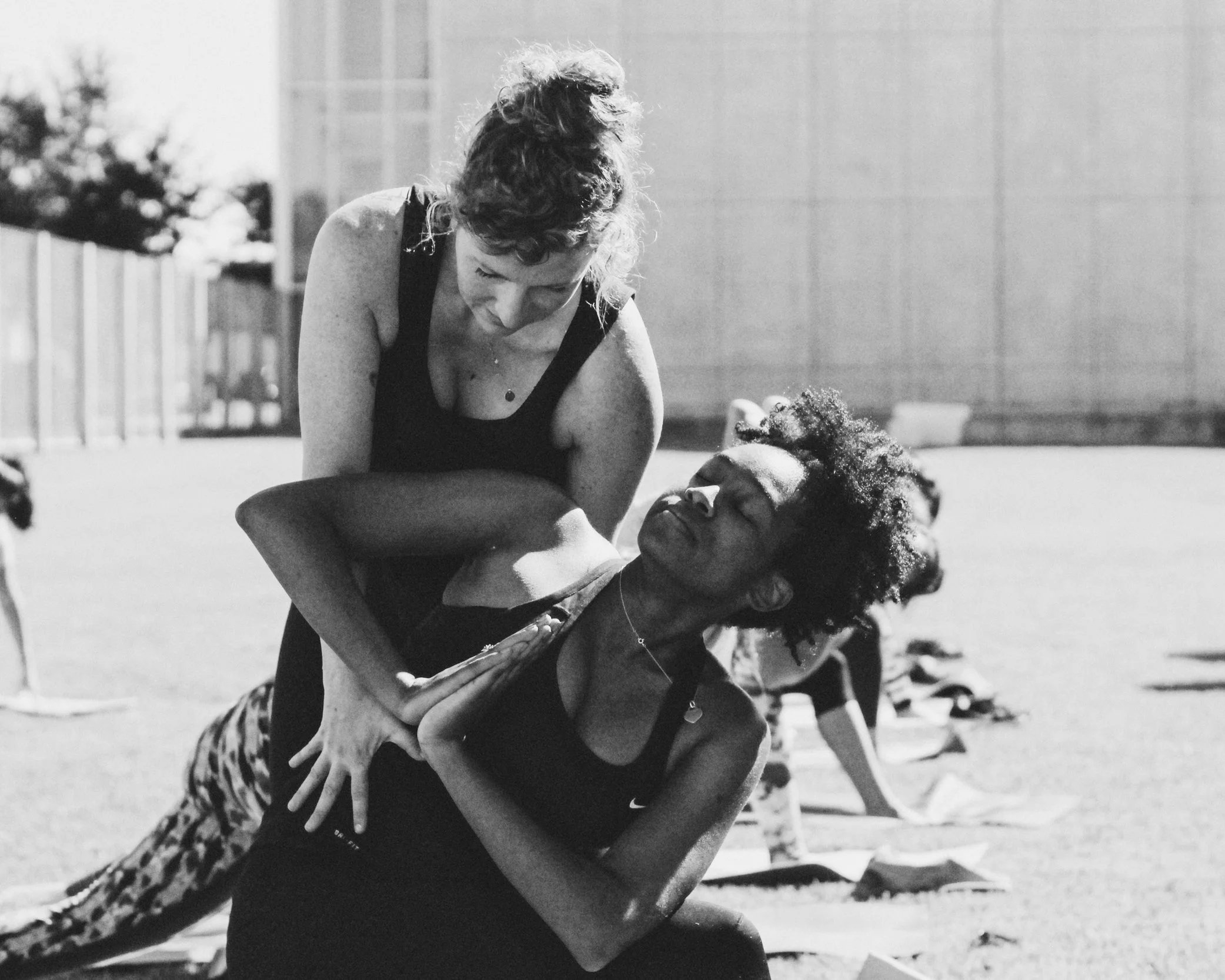 Two women practicing yoga outdoors, one assisting the other with a pose on a grassy field with mats and other people in the background.