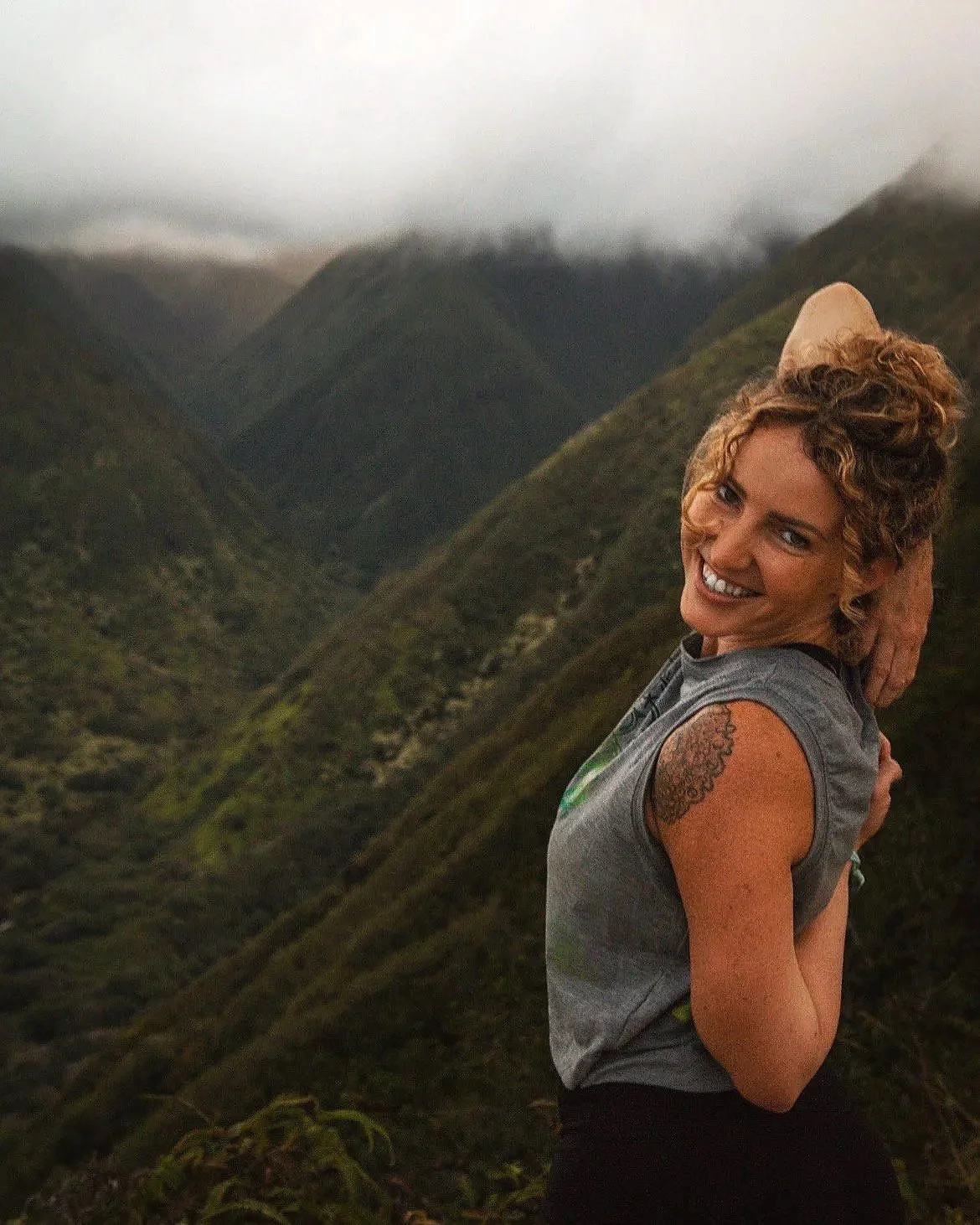 Woman with curly hair smiling and posing outdoors with green mountains in the background, wearing a gray sleeveless shirt and a tan hat.