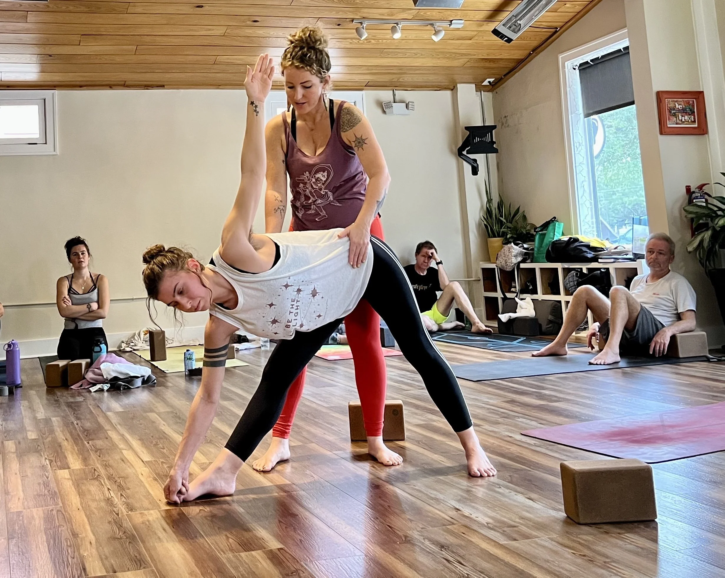 A yoga class in progress with an instructor assisting a woman in a white shirt and black leggings into a forward bend pose. Several students are seated or lying on yoga mats in a wooden-floored studio with natural light. Some students are watching, others are resting or preparing for poses.