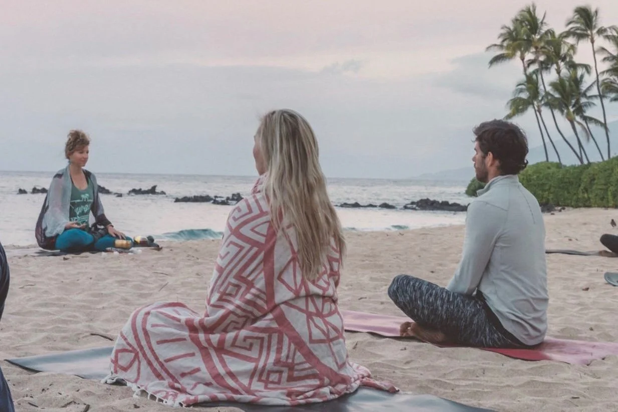 Three people sitting on the sandy beach facing a woman who is meditating or practicing yoga near the ocean. The beach has palm trees and rocks in the background, with a cloudy sky.