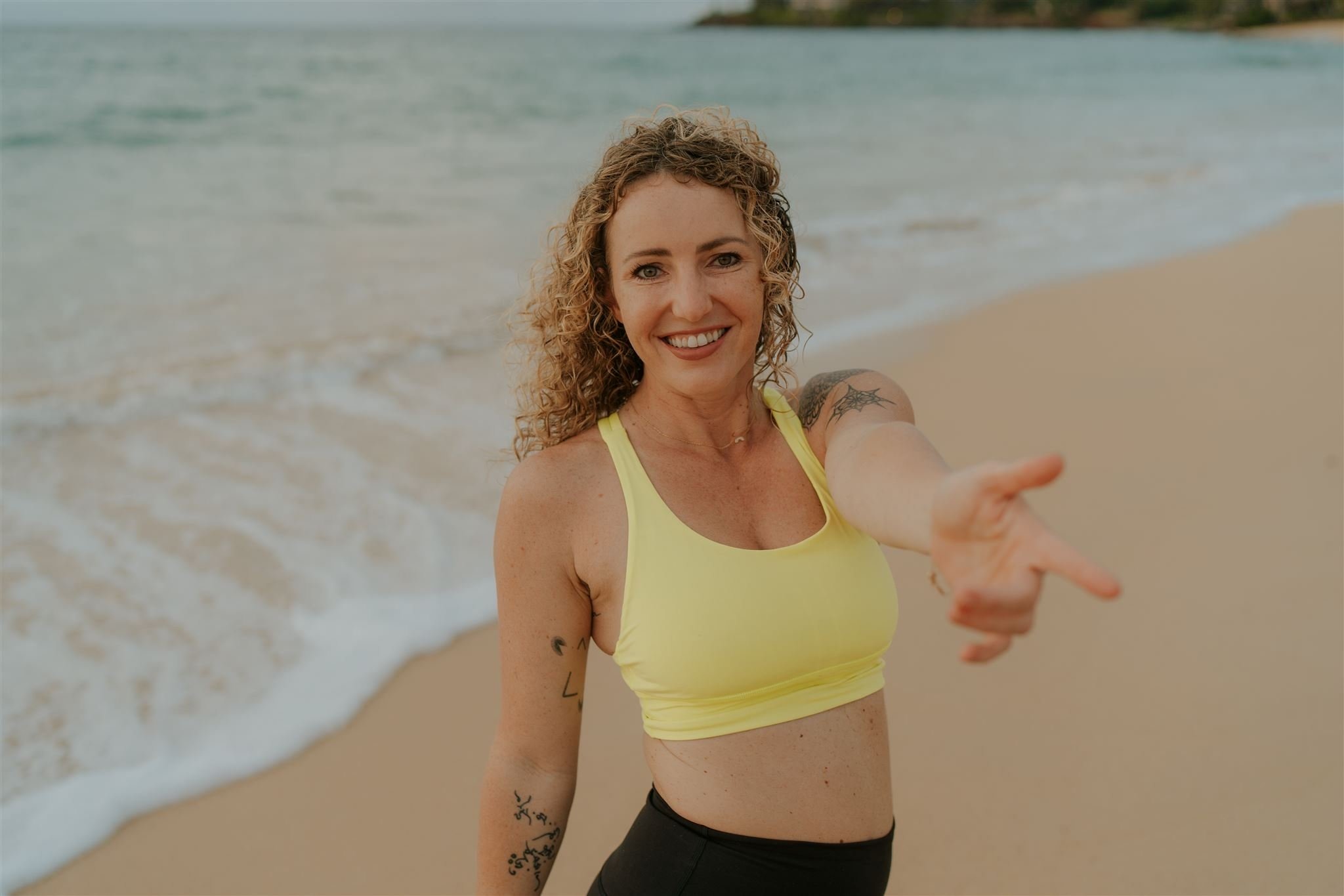 A woman with curly hair smiling on the beach, wearing a yellow sports bra and black shorts, reaching out towards the camera.