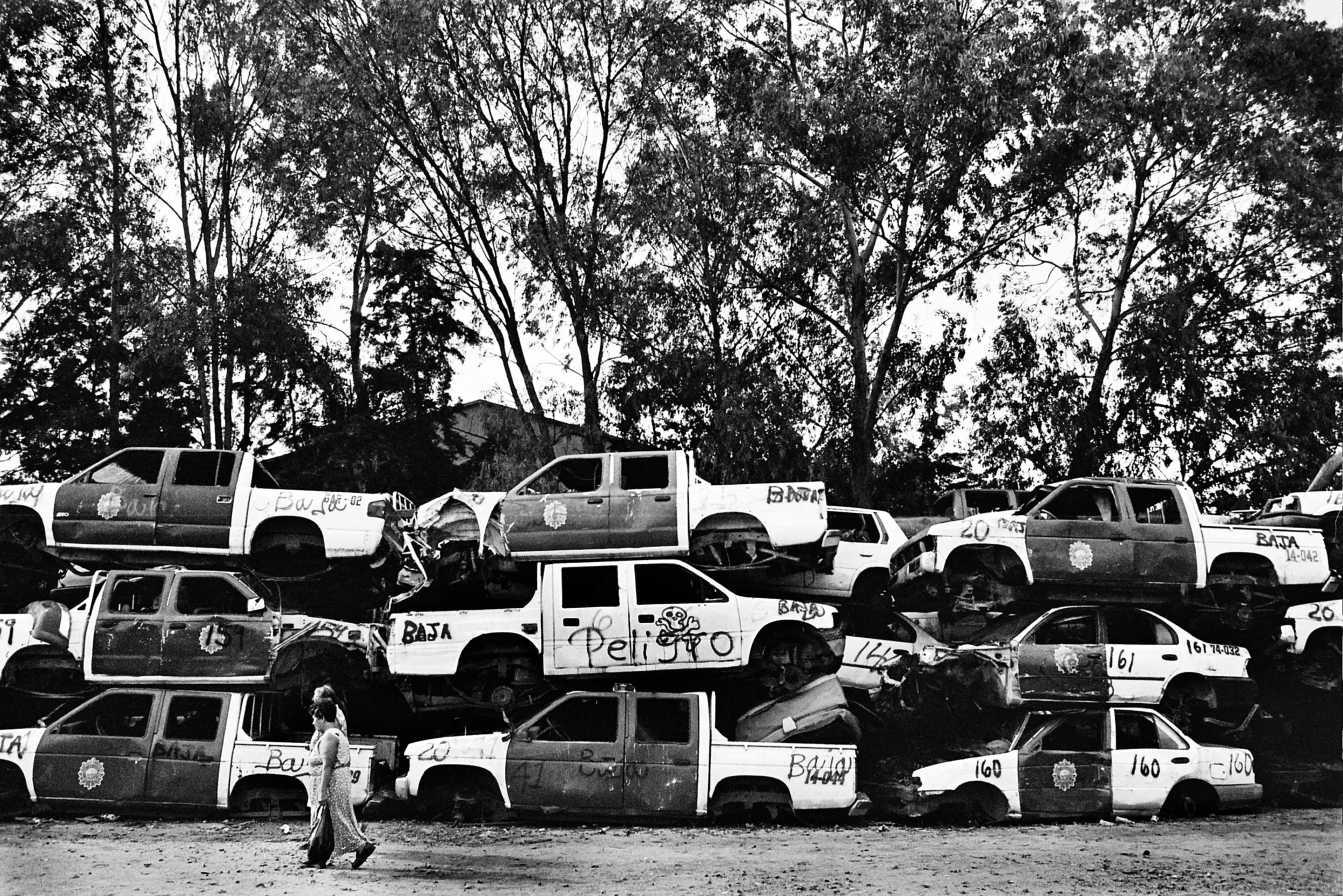  Women walk by a wall of wrecked police trucks. 