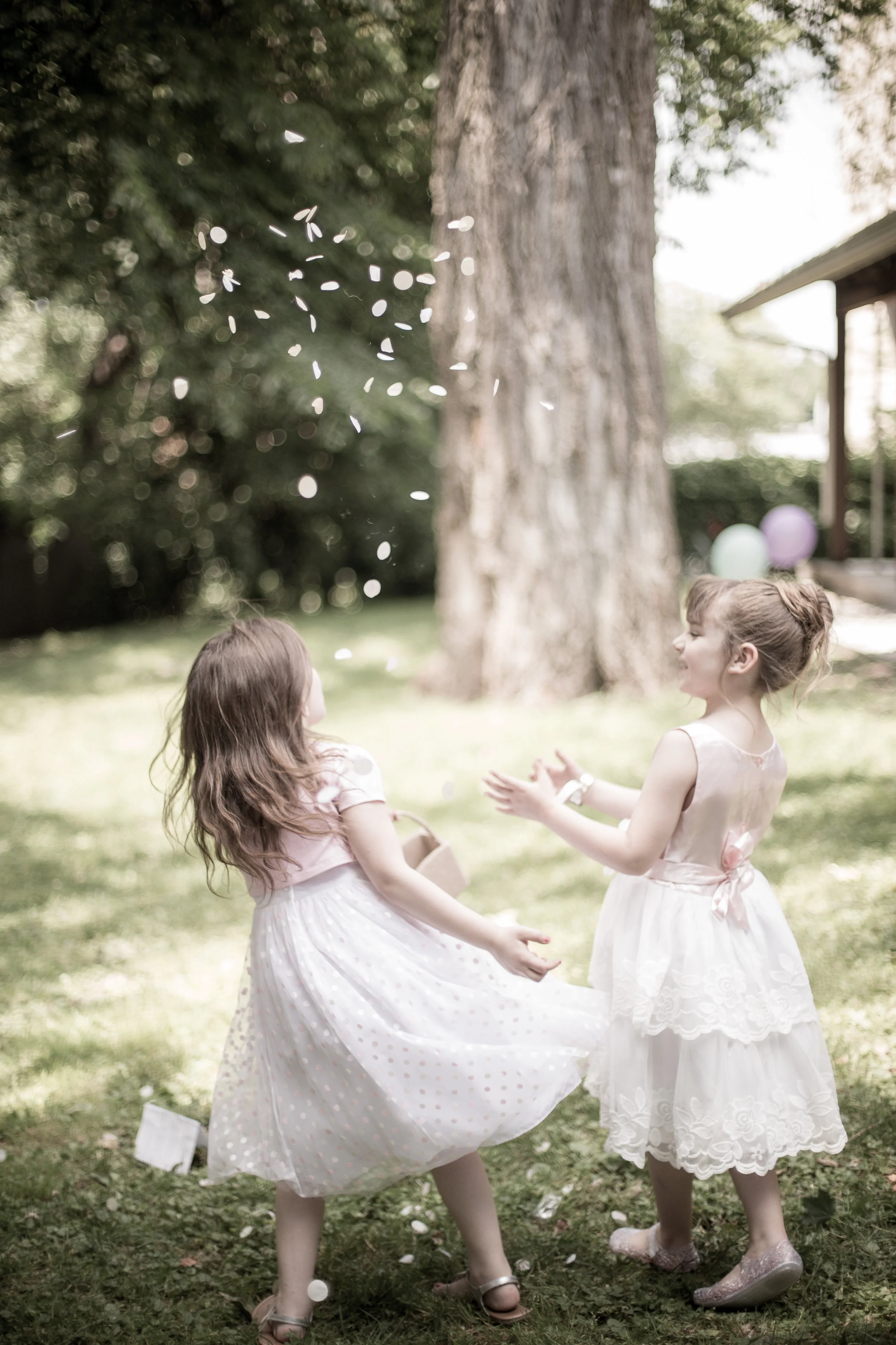 Two young girls in white dresses playing outside in a grassy area with balloons and a large tree in the background.
