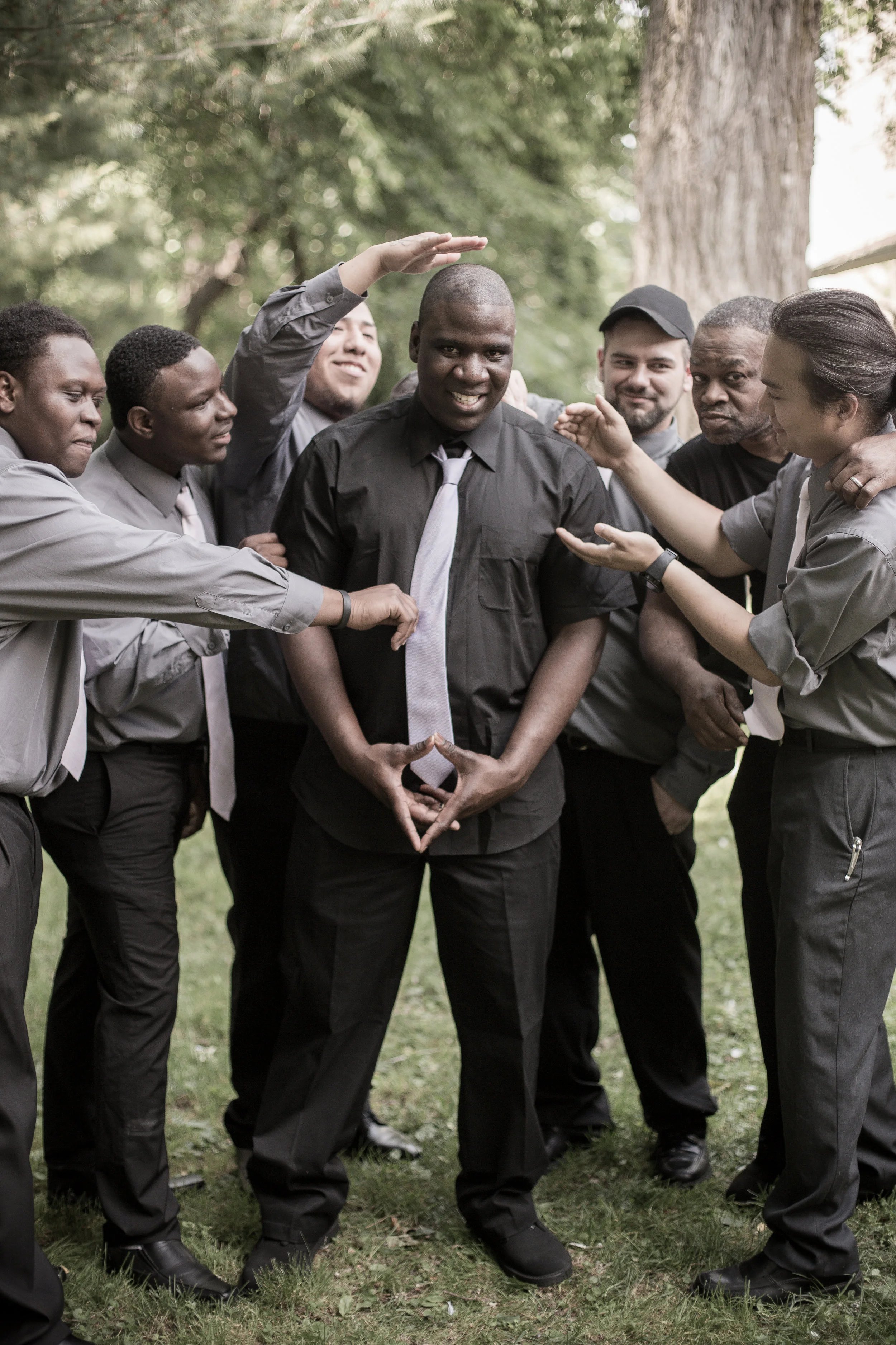 A group of diverse people in black and gray clothing forming a circle outdoors, with a young man in the center making a hand gesture, and others touching his shoulders and head, celebrating or engaging in a team activity.