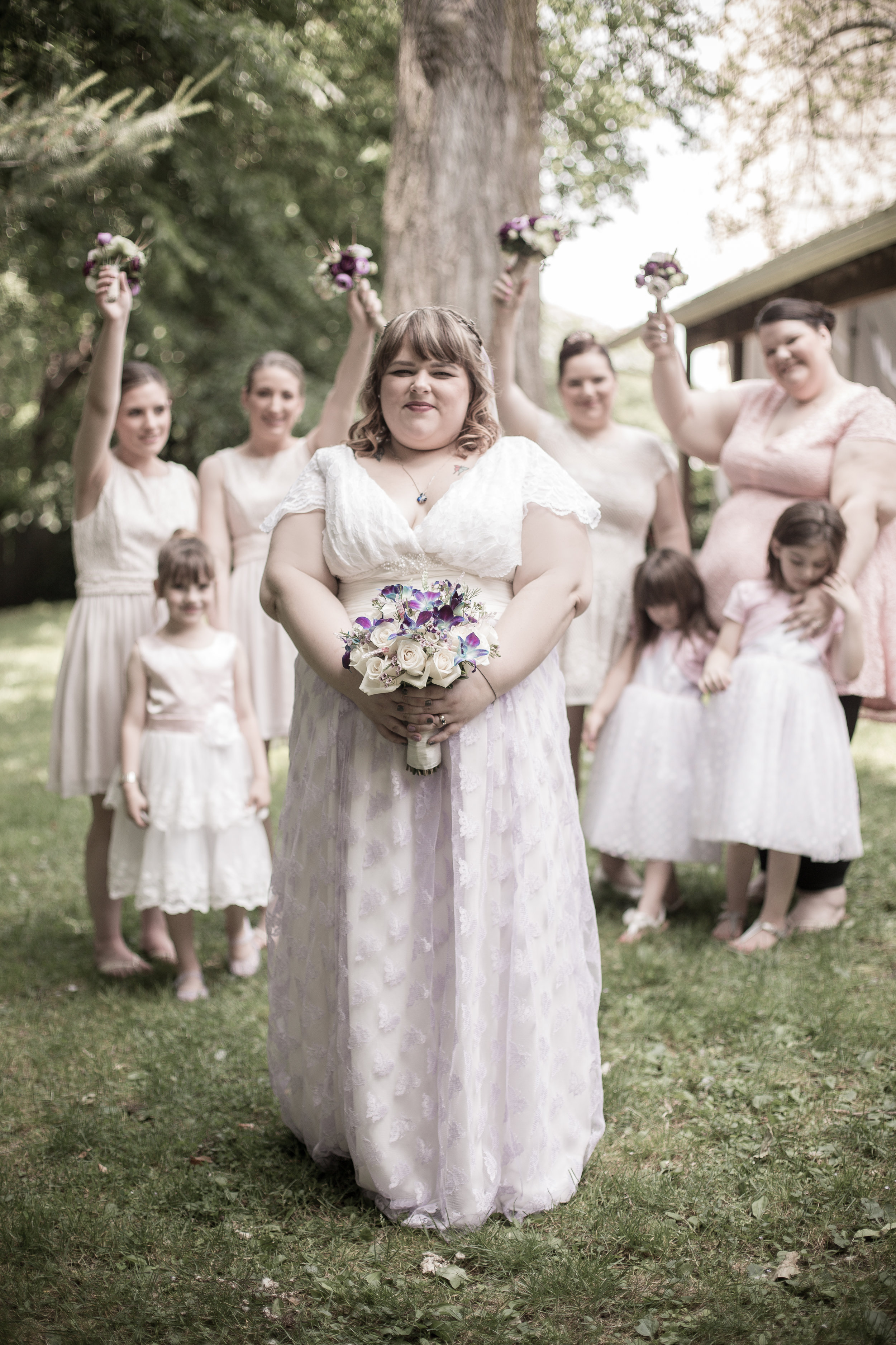 A bride holding a bouquet of white and purple flowers stands outdoors, surrounded by bridesmaids and flower girls in light-colored dresses, with trees and a wooden structure in the background.