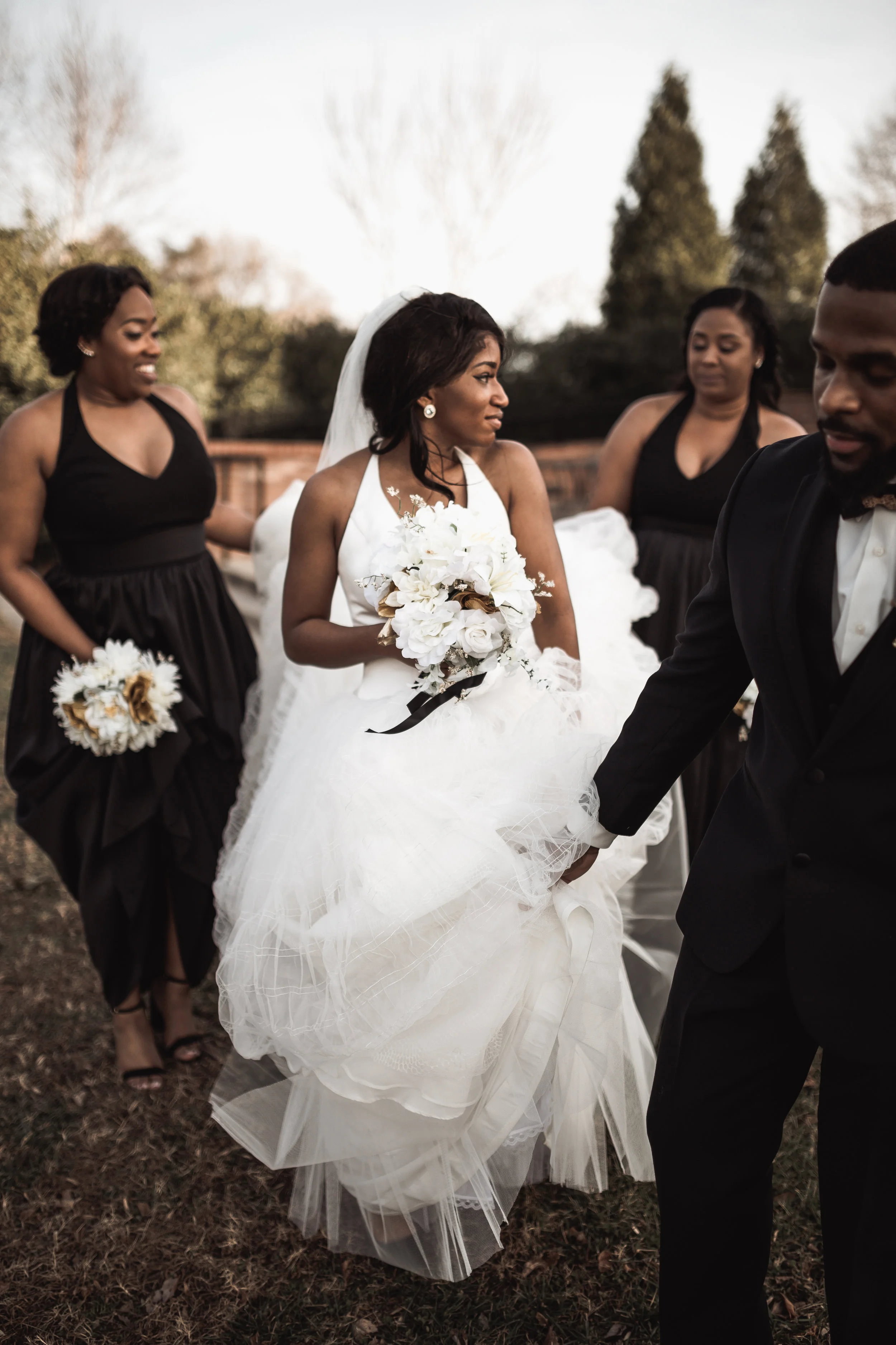 A bride in a white wedding gown holding a bouquet, surrounded by bridesmaids and groomsmen, outdoors on a cloudy day with trees in the background.