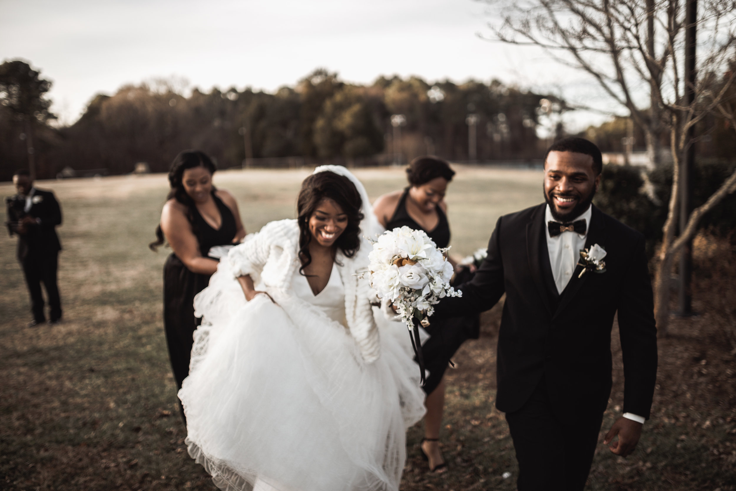 A happy bride and groom walking outdoors with friends on their wedding day, holding a bouquet of white flowers, with friends smiling in the background.
