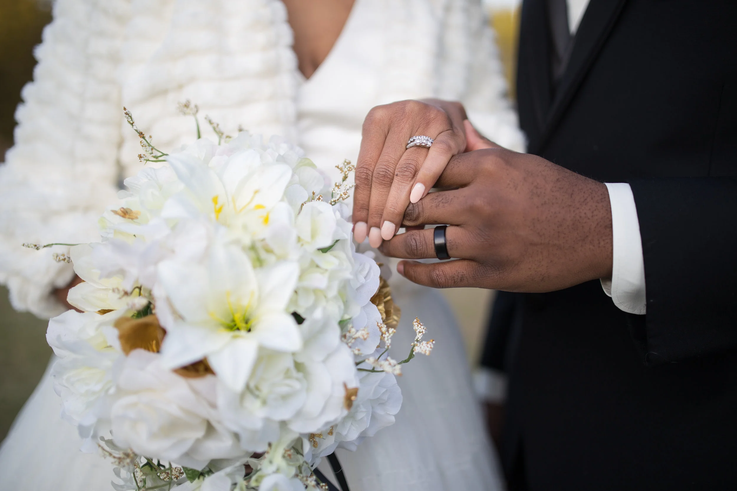 Close-up of a newlywed couple holding hands during a wedding ceremony, with focus on their wedding rings, white floral bouquet in the foreground, and they dressed in formal wedding attire.