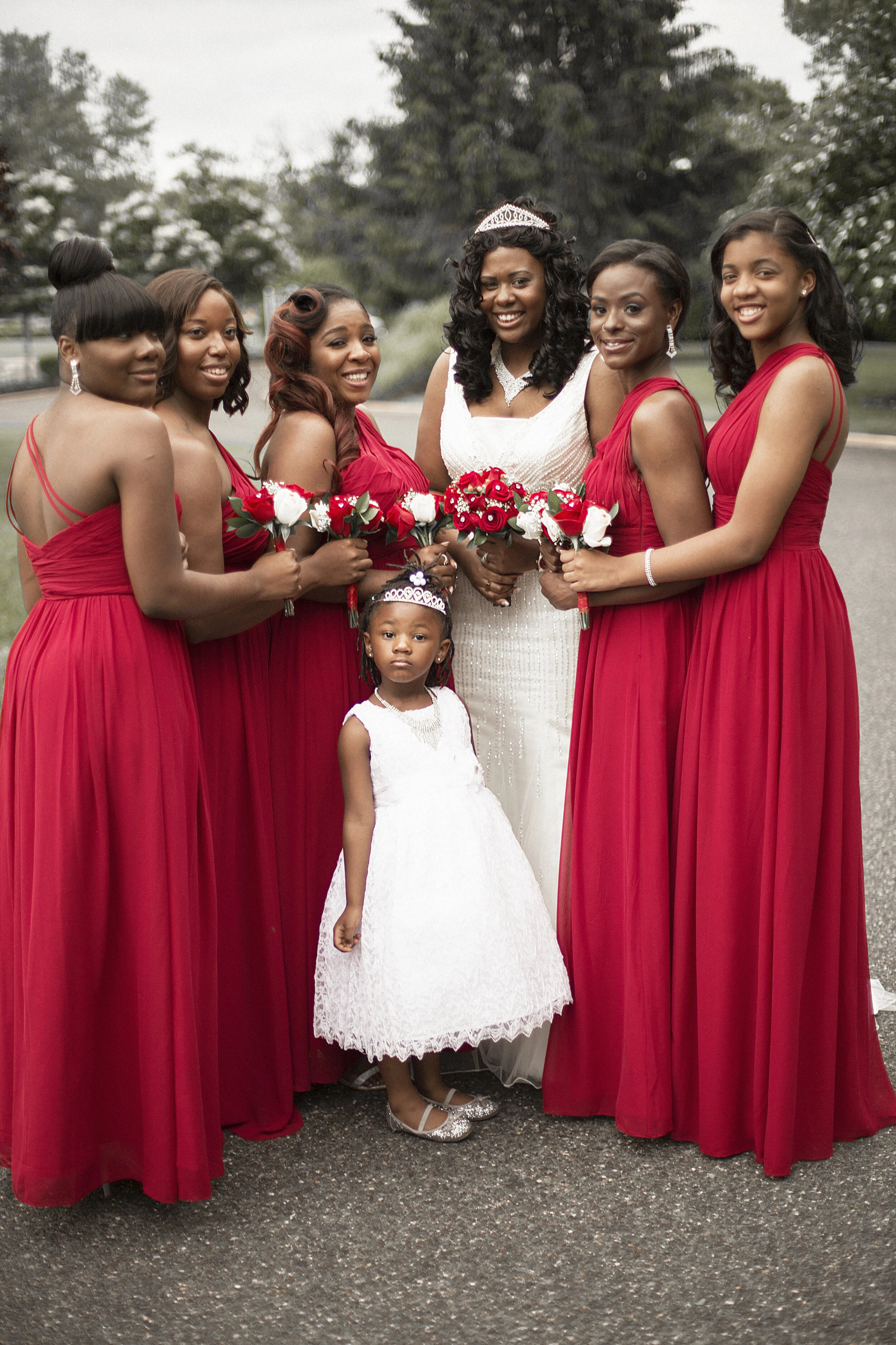 A group of women in red dresses and two young girls in white dresses, with the women holding bouquets and one woman wearing a tiara, standing outdoors on a paved area with trees in the background, celebrating a special occasion.