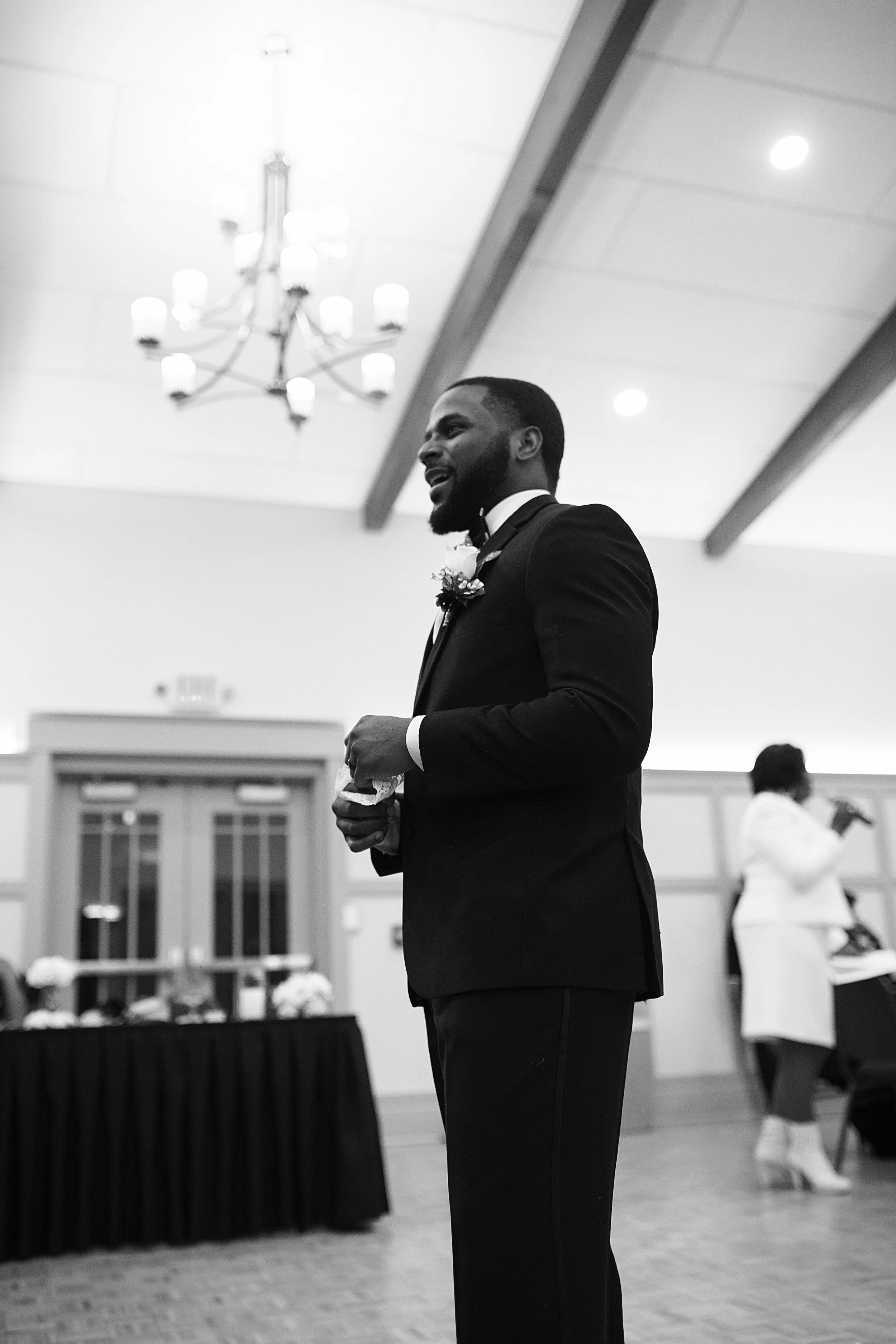 A man in a tuxedo with a boutonniere standing in a banquet hall with a chandelier hanging from the ceiling.