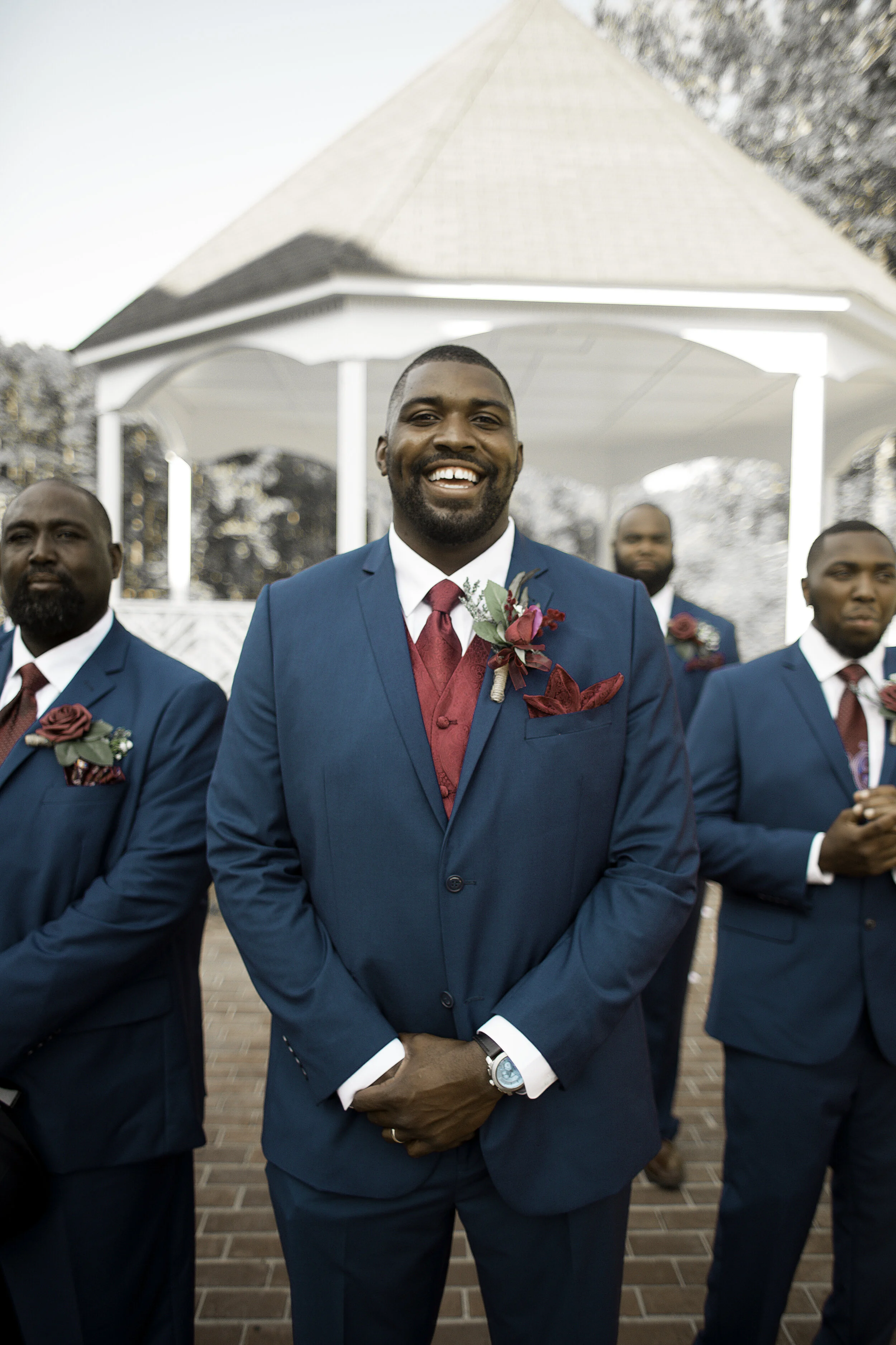 A group of men dressed in formal teal suits with burgundy accents, standing outdoors on a brick patio in front of a white gazebo. The man in the center, smiling and wearing a watch, has a boutonniere on his lapel.