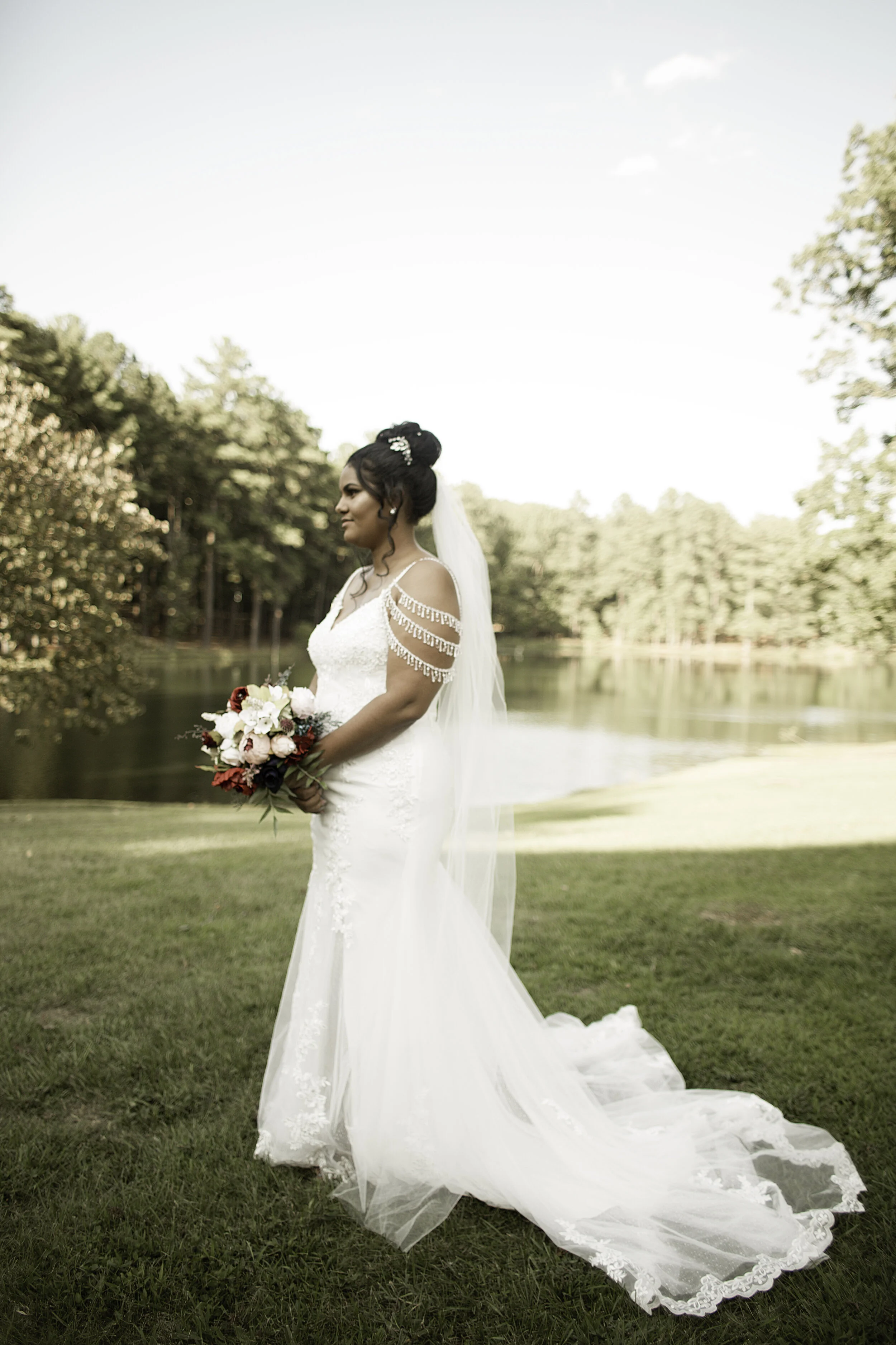 A bride in a white wedding gown with lace details holding a bouquet of flowers standing on a grassy area near a lake with trees and a partly cloudy sky in the background.