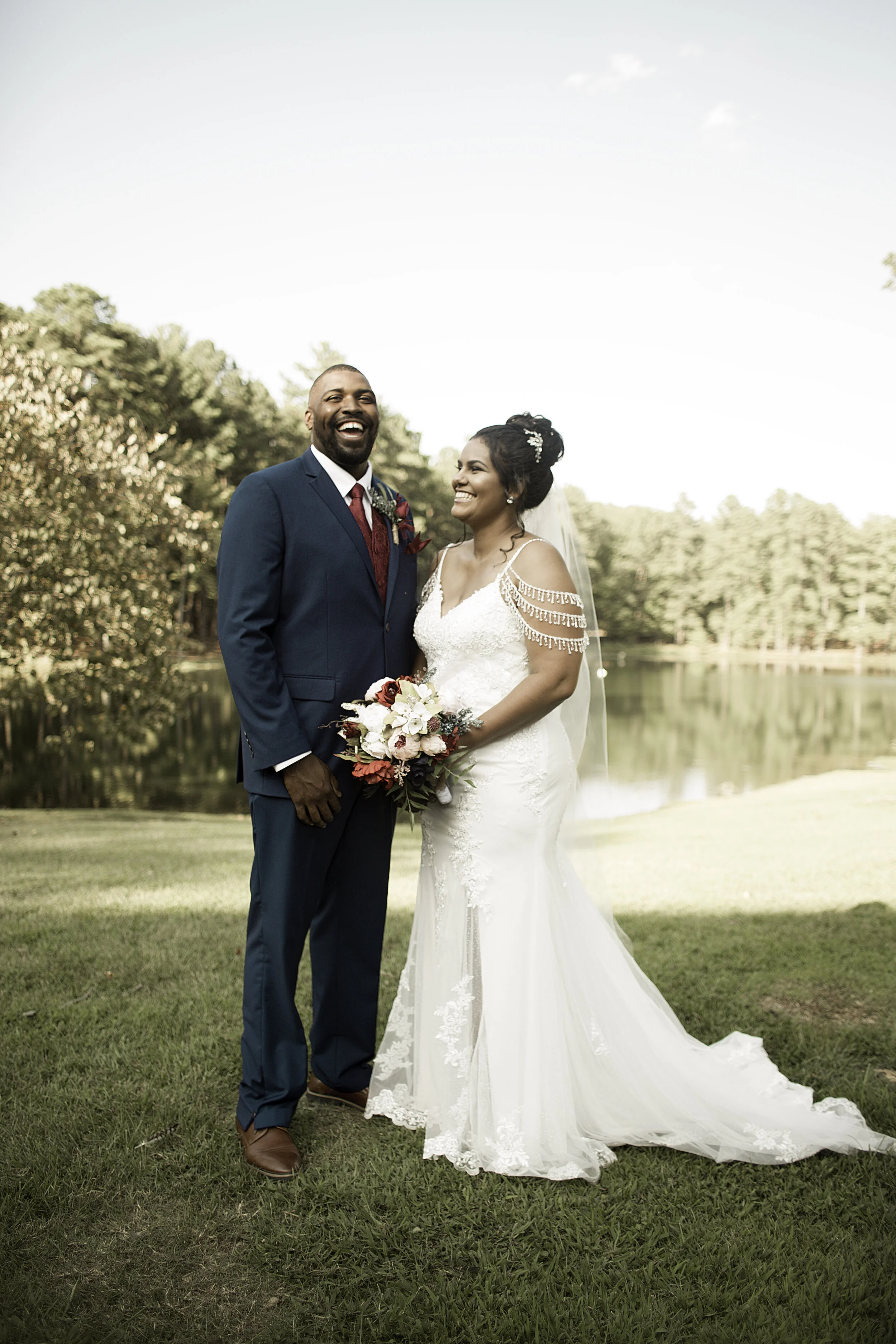 A happy bride and groom standing outdoors by a lake, smiling, with the bride holding a bouquet of flowers, trees, and a lake in the background.