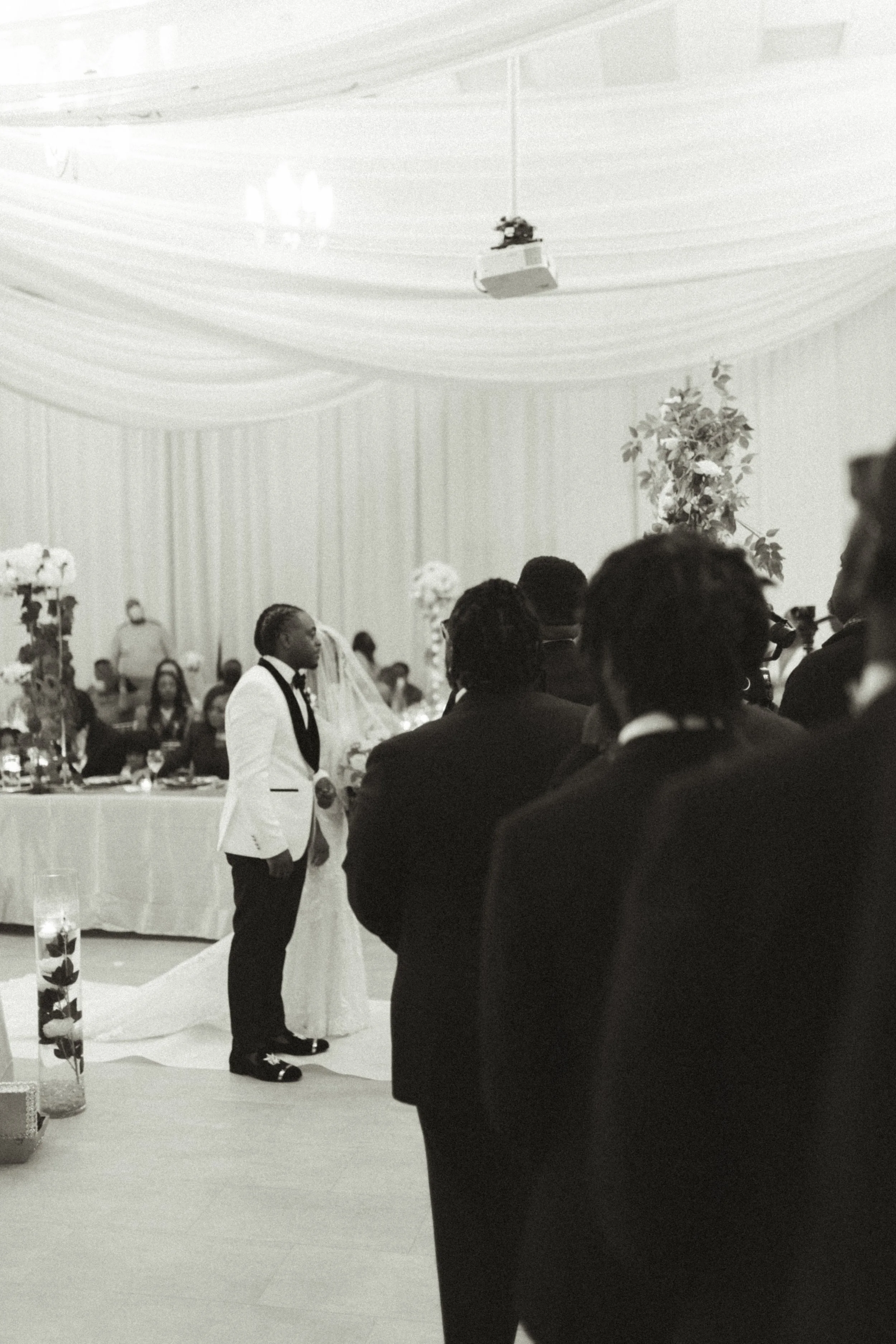 A black and white photo of a wedding reception with a bride and groom standing together in the background. The bride is wearing a wedding gown, and the groom is dressed in a suit. Several guests are seated and standing around, observing the ceremony.