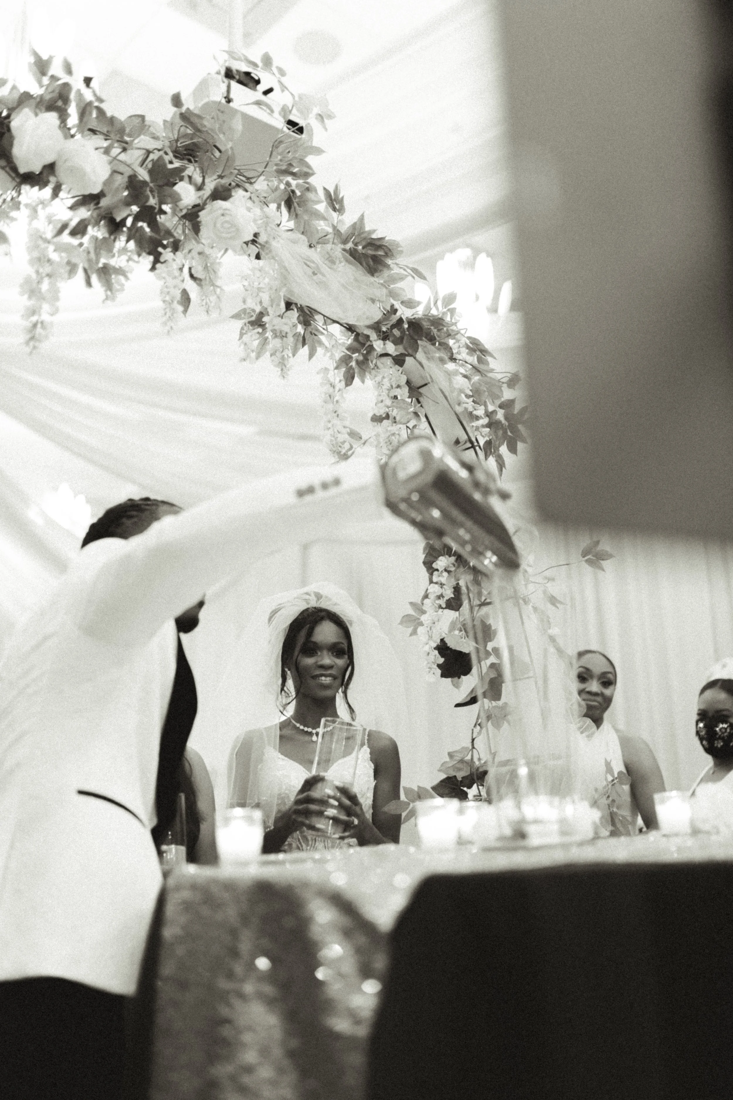 Black and white photo of a wedding reception with women in dresses, some holding drinks, under floral decorations and a draped ceiling.