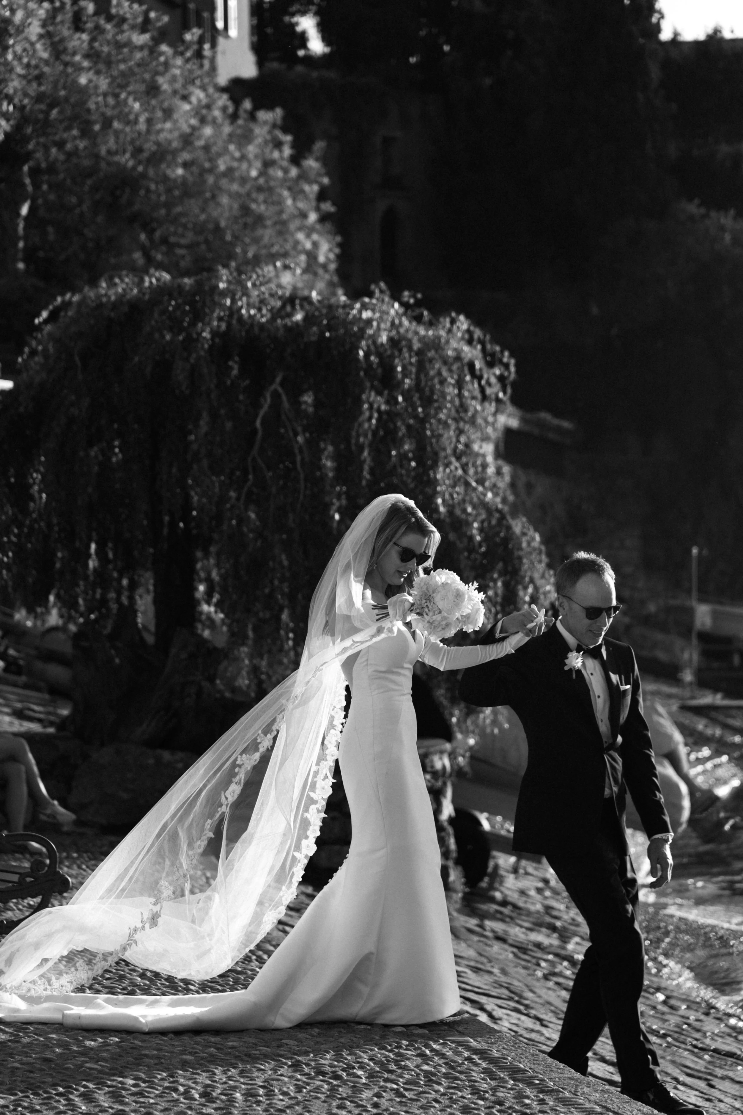 A bride in a white wedding dress with a veil and sunglasses holding a bouquet walking with a groom in a tuxedo on a cobblestone street outdoors, surrounded by trees and people.
