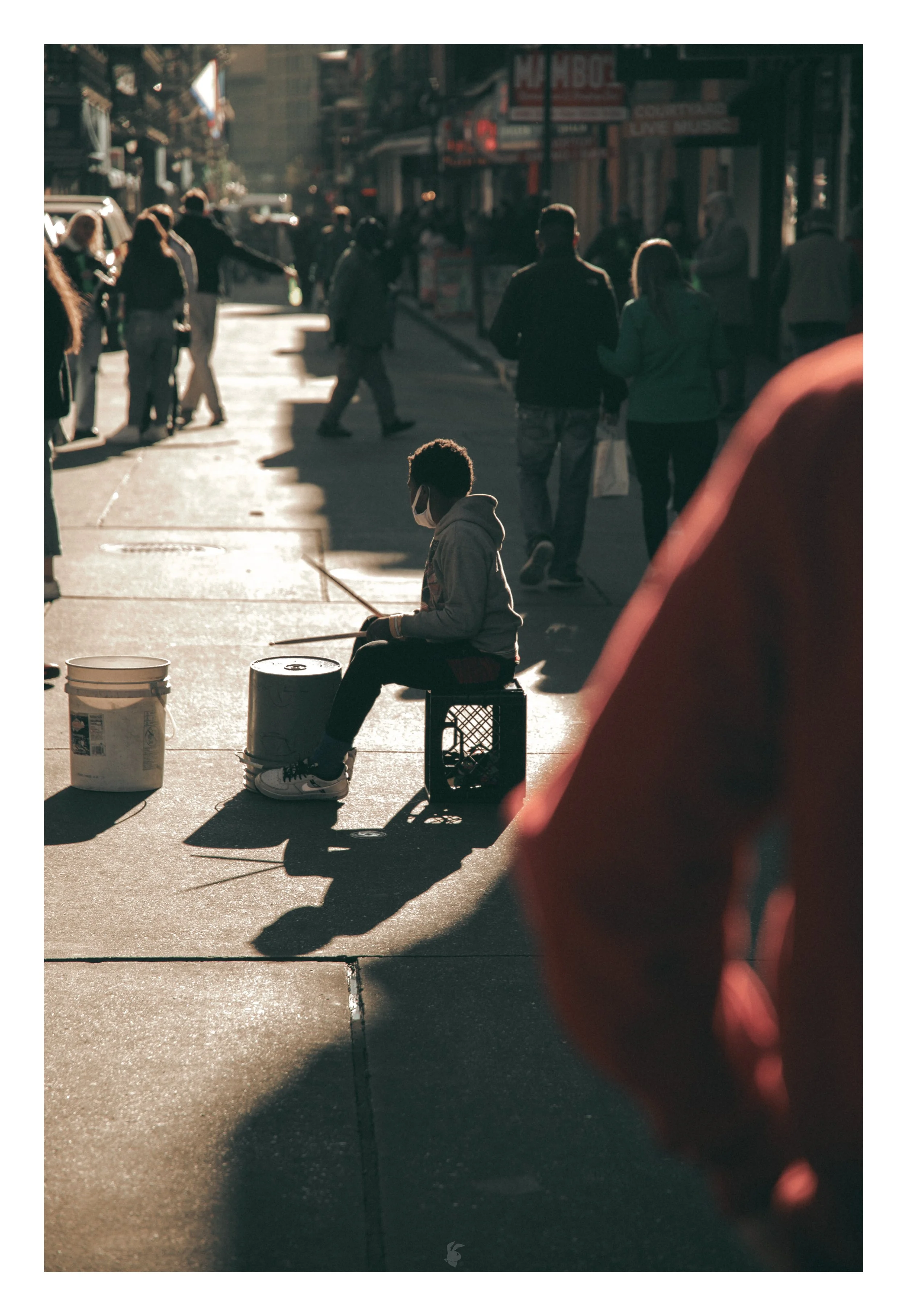 A boy sitting on a crate playing drums on a city sidewalk during sunset, with pedestrians walking by in the background.