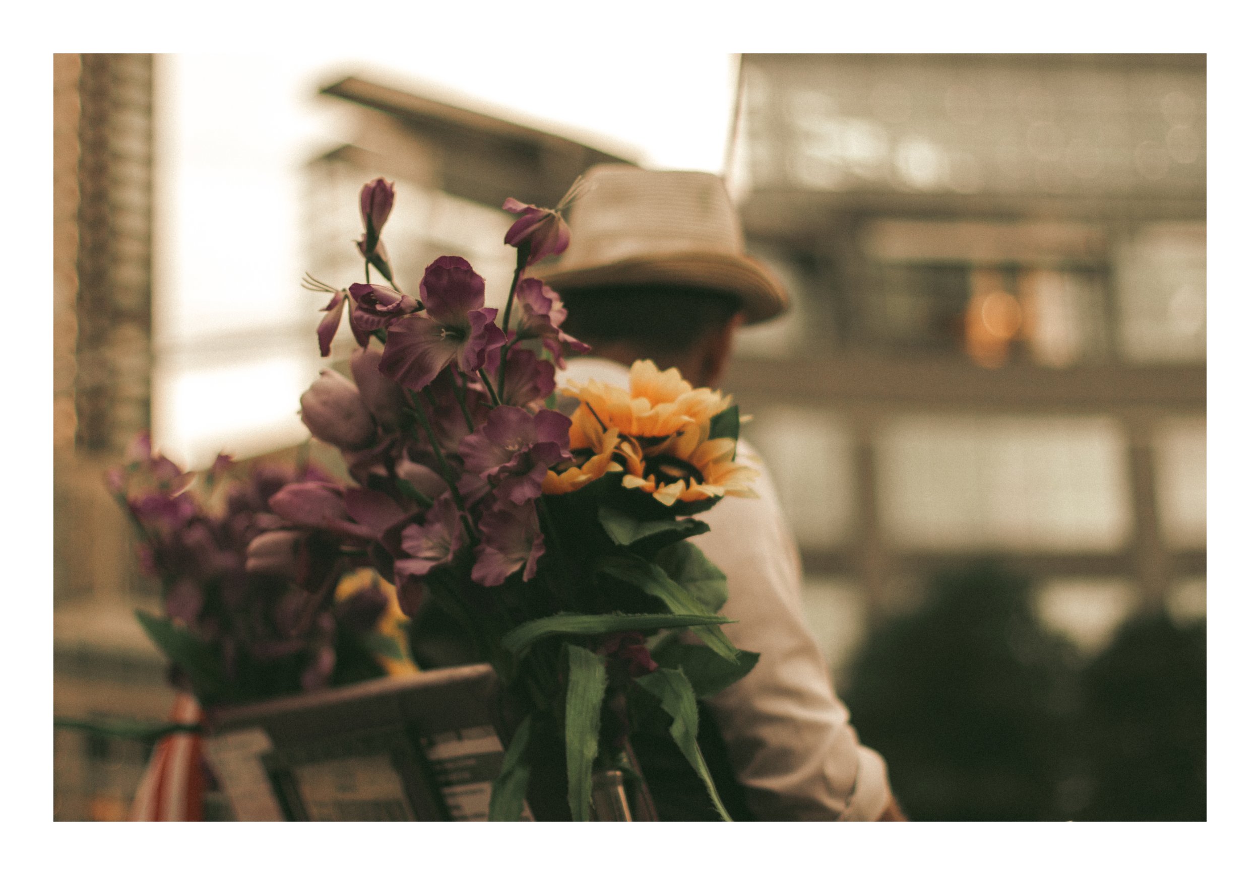 A person wearing a hat sits in a cozy indoor space, with a bouquet of pink and yellow flowers in front of them, and appear to be reading a book.