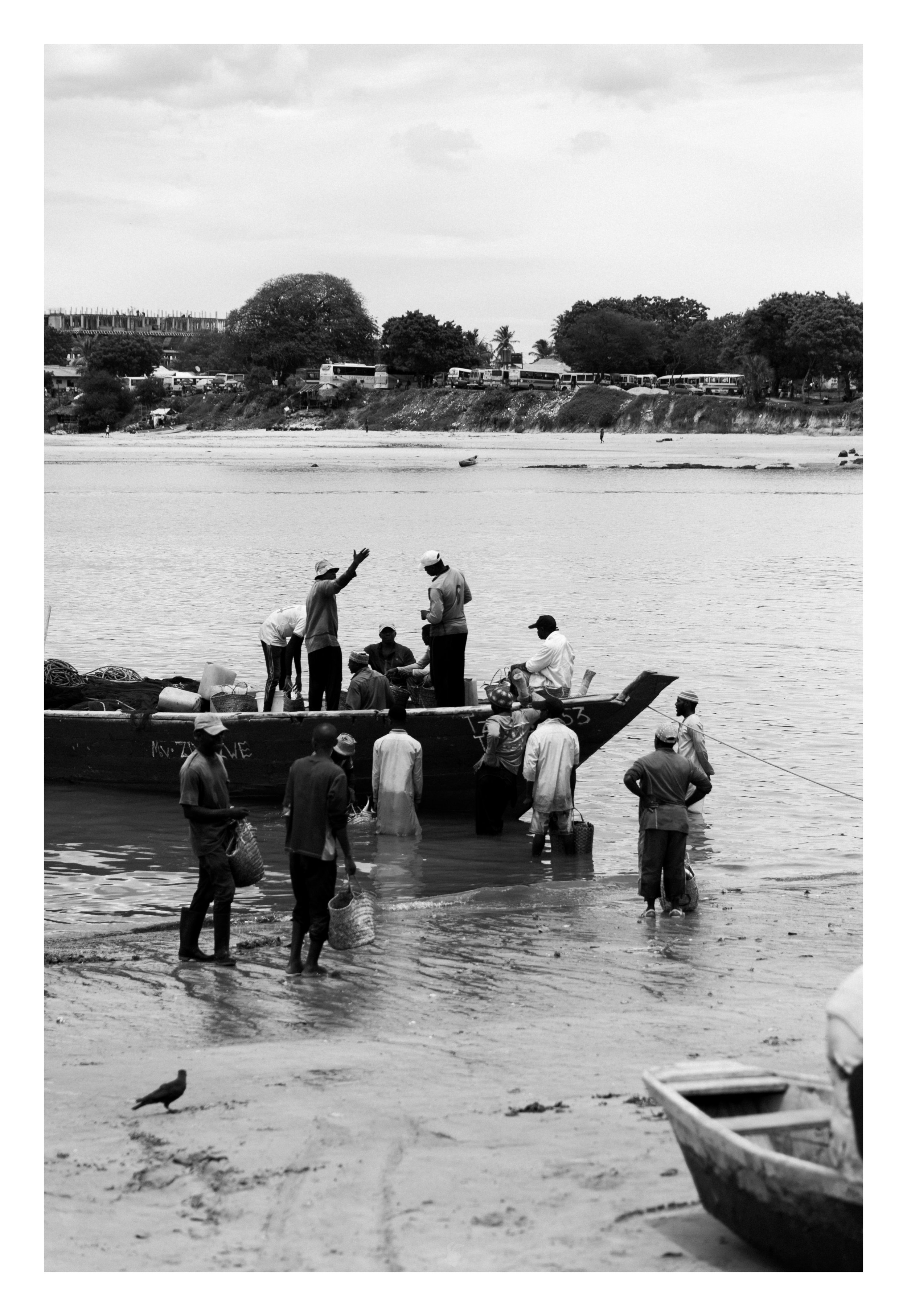 People gathering at a riverbank, some standing in shallow water, others on the shore, with boats in the foreground and a distant shoreline with trees and vehicles in the background.