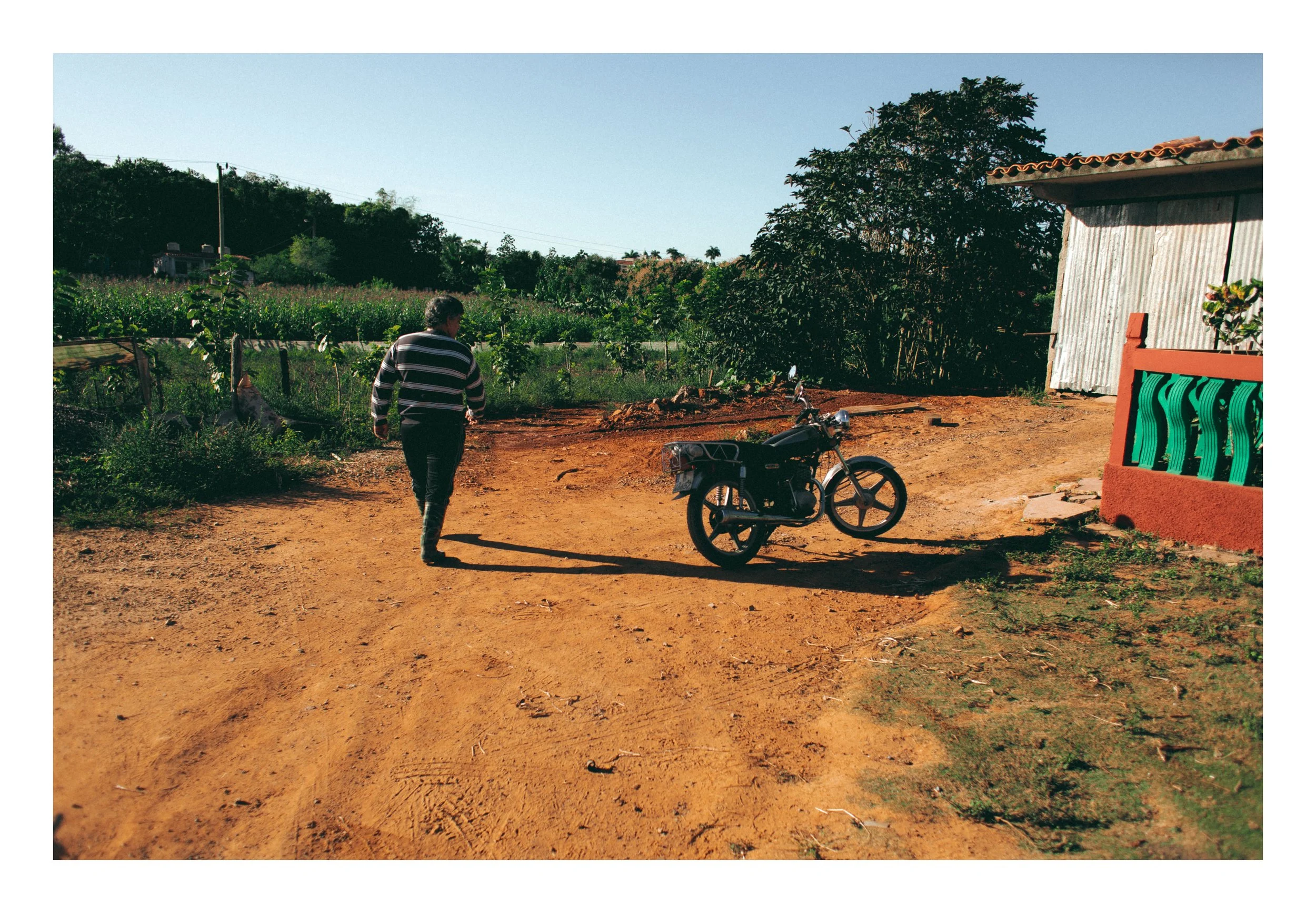 A person walking on a dirt road next to a parked motorcycle, with a building and green trees in the background under a clear blue sky.