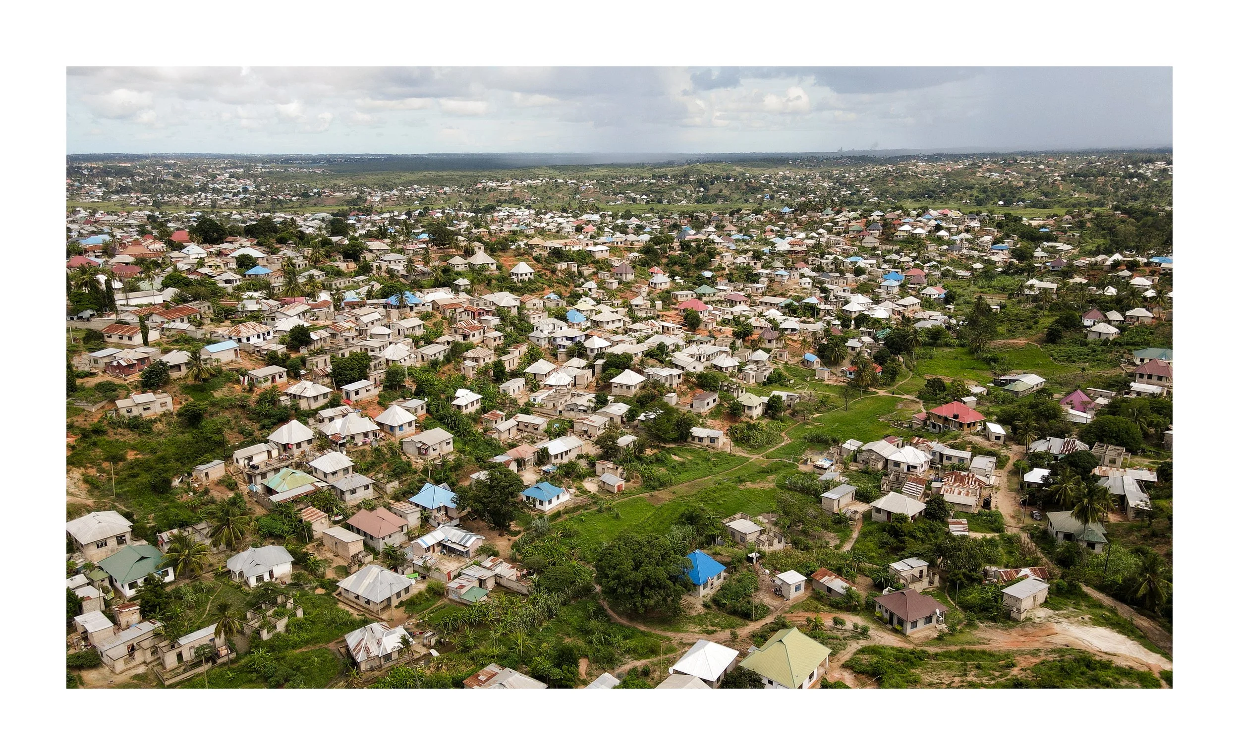 Aerial view of a densely populated residential area with numerous small houses, many with colorful roofs, scattered among green trees and vegetation, extending into the horizon under a cloudy sky.
