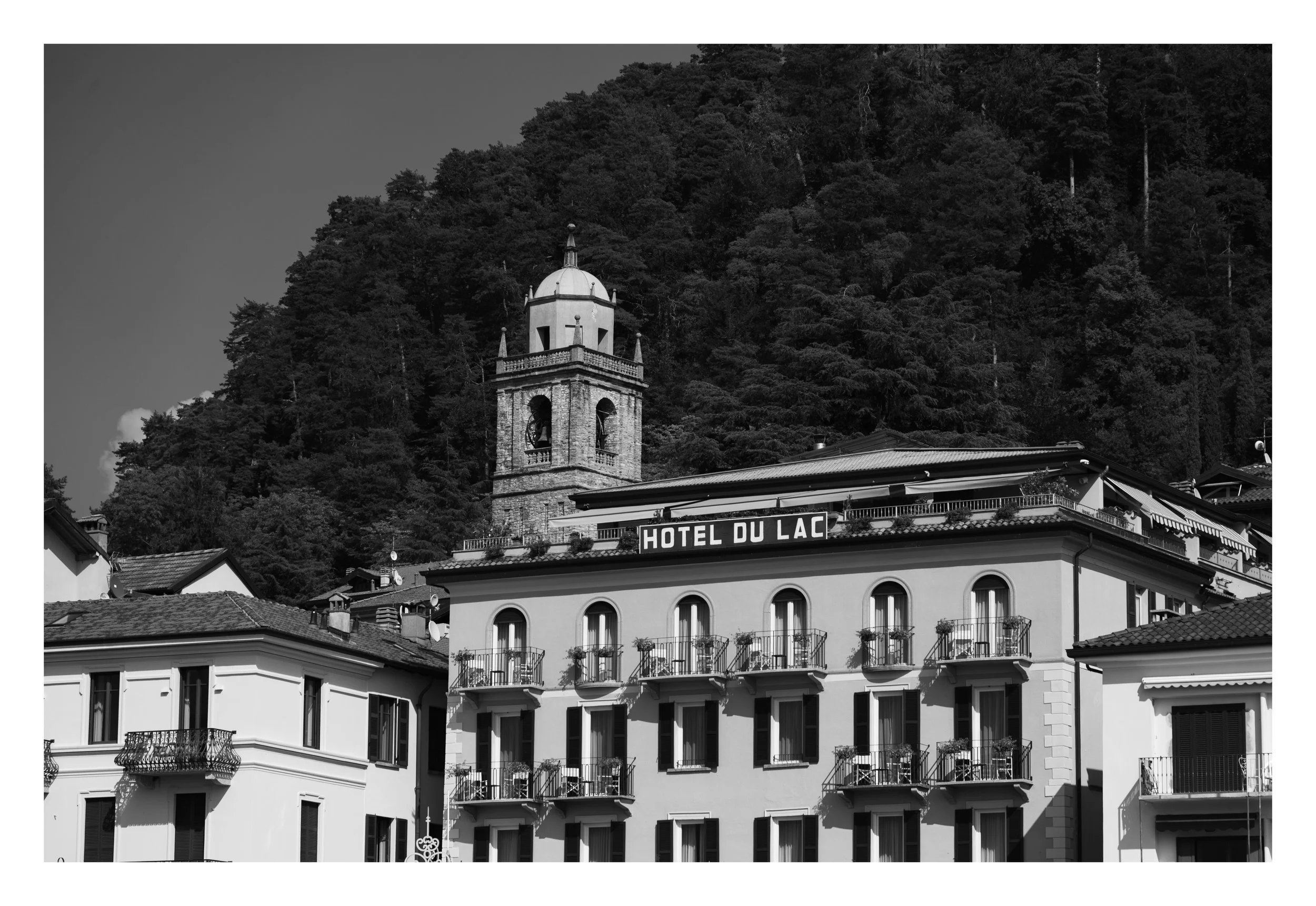 Black and white photo of a hillside with trees, a bell tower, and a building with a sign that reads 'Hotel du Lac' and multiple balconies.