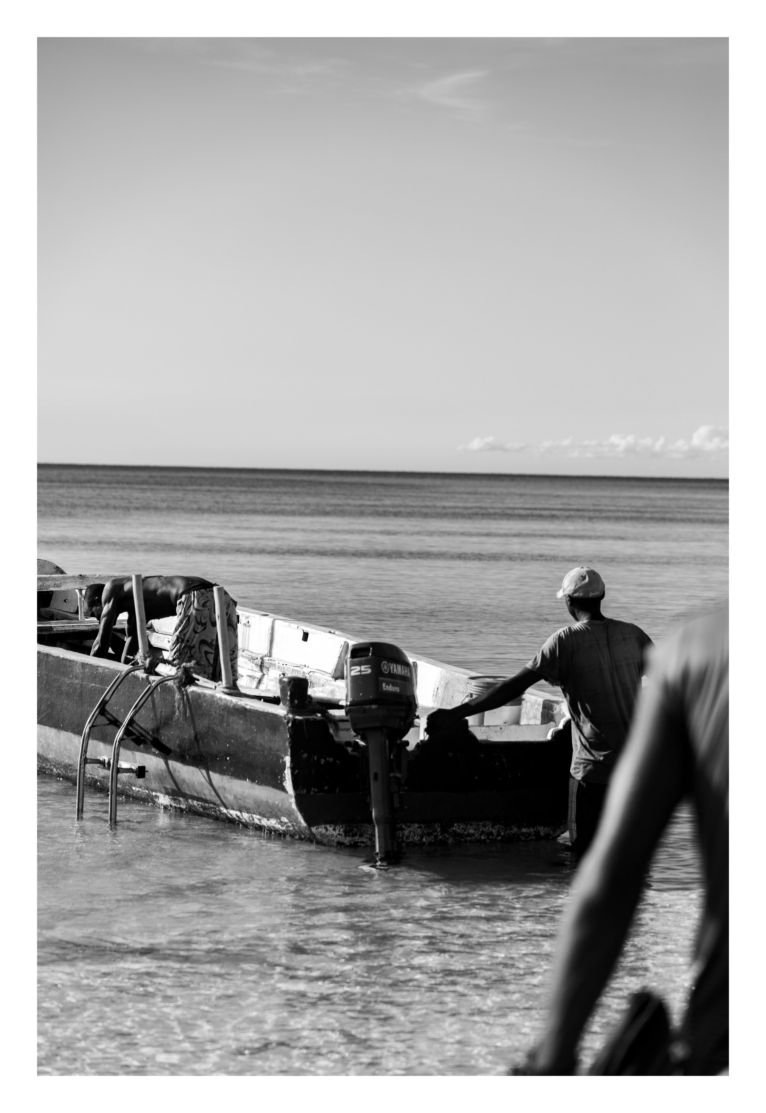 Two men are near a boat on the water, one is working on the boat while a third person, wearing a cap, stands in the foreground. The water and sky are visible in the background.