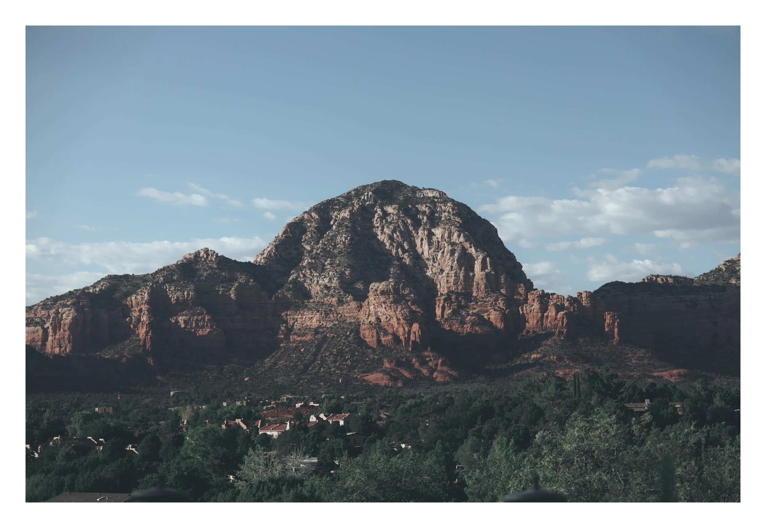 A large mountain with a layered, rugged appearance under a bright blue sky with scattered clouds, with a residential area and trees in the foreground.