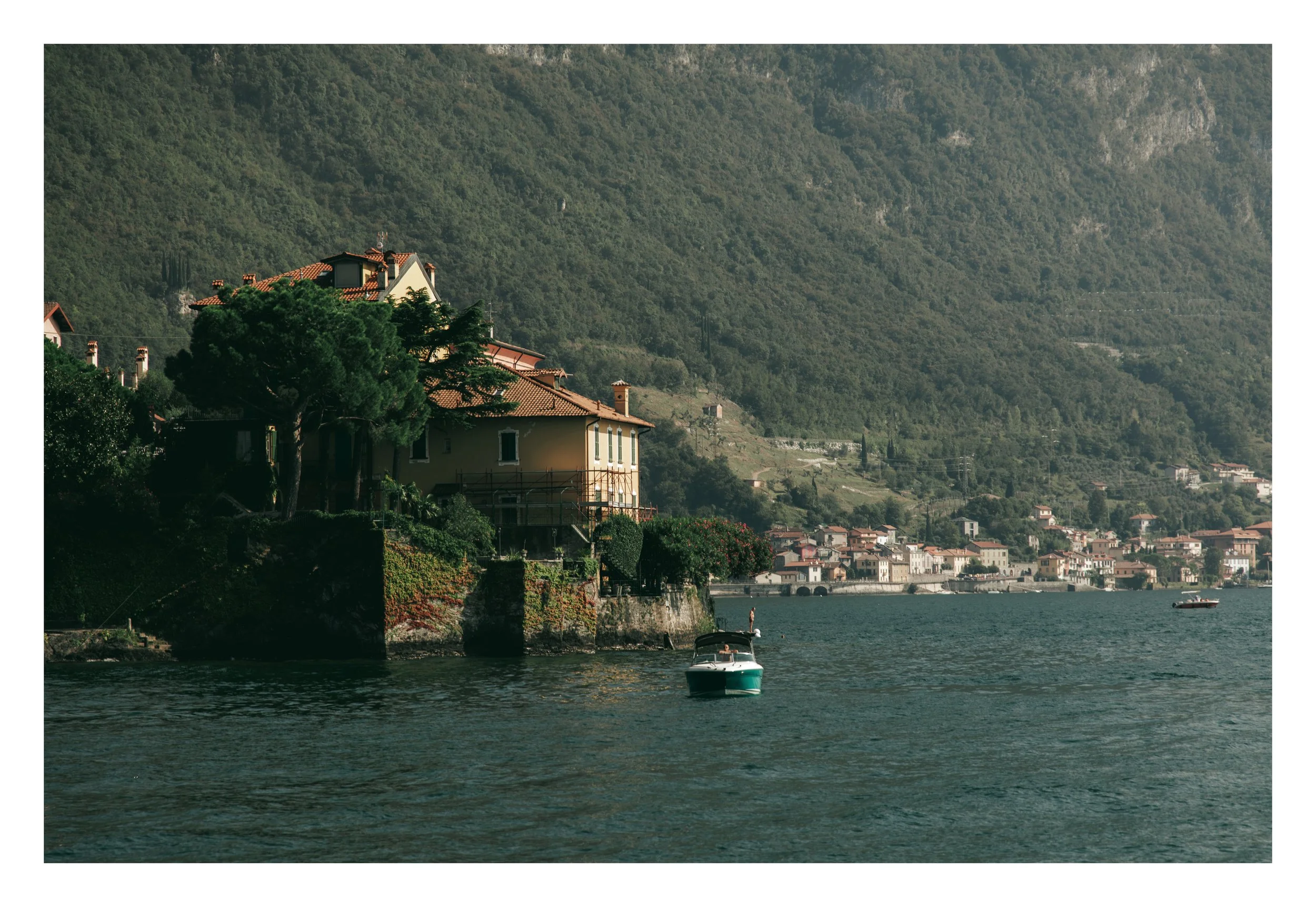 A lakeside town with houses and lush green mountains in the background, with boats on the water.