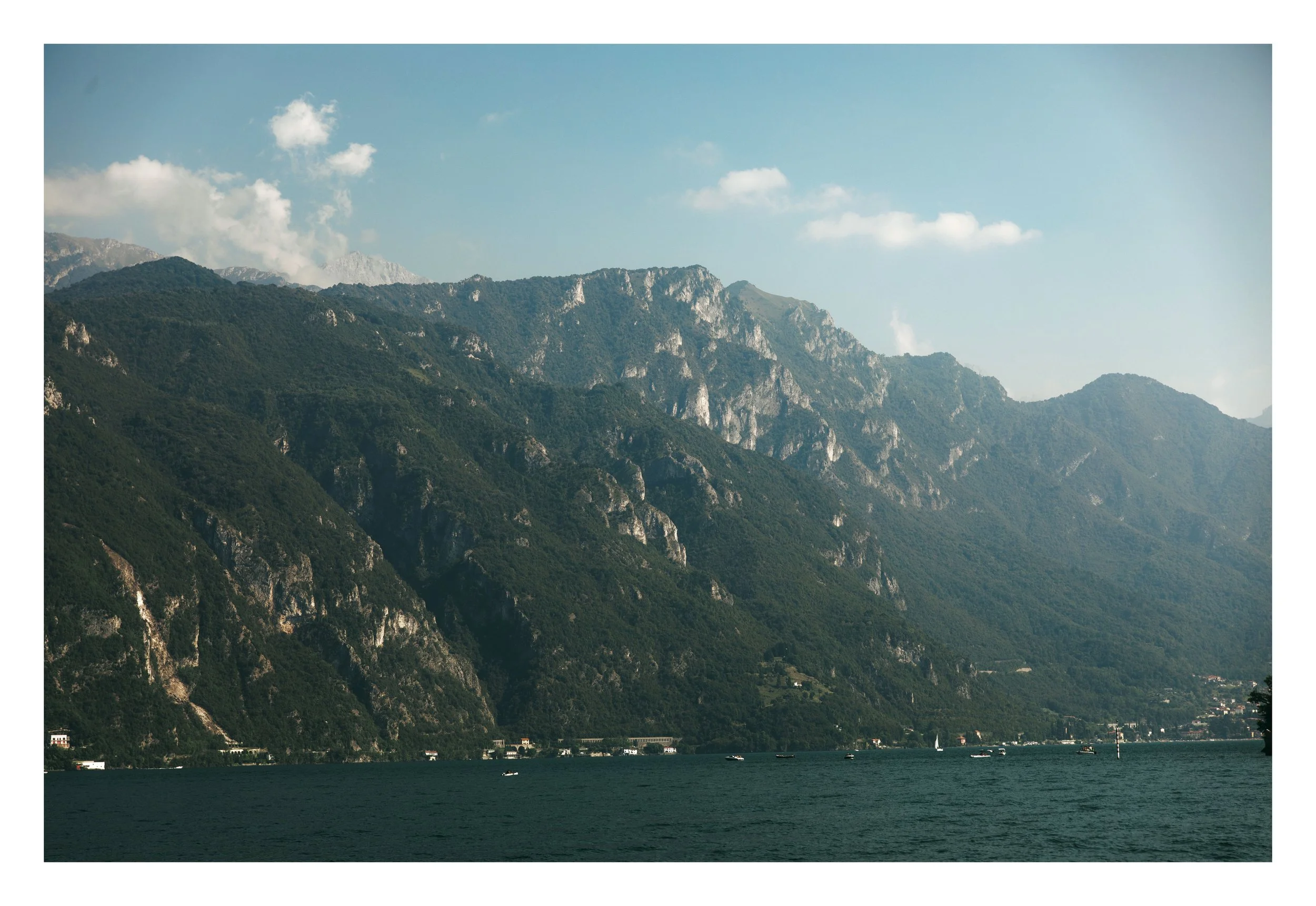 Mountains with lush green trees and rocky cliffs, near a body of water with boats, under a blue sky with scattered clouds.