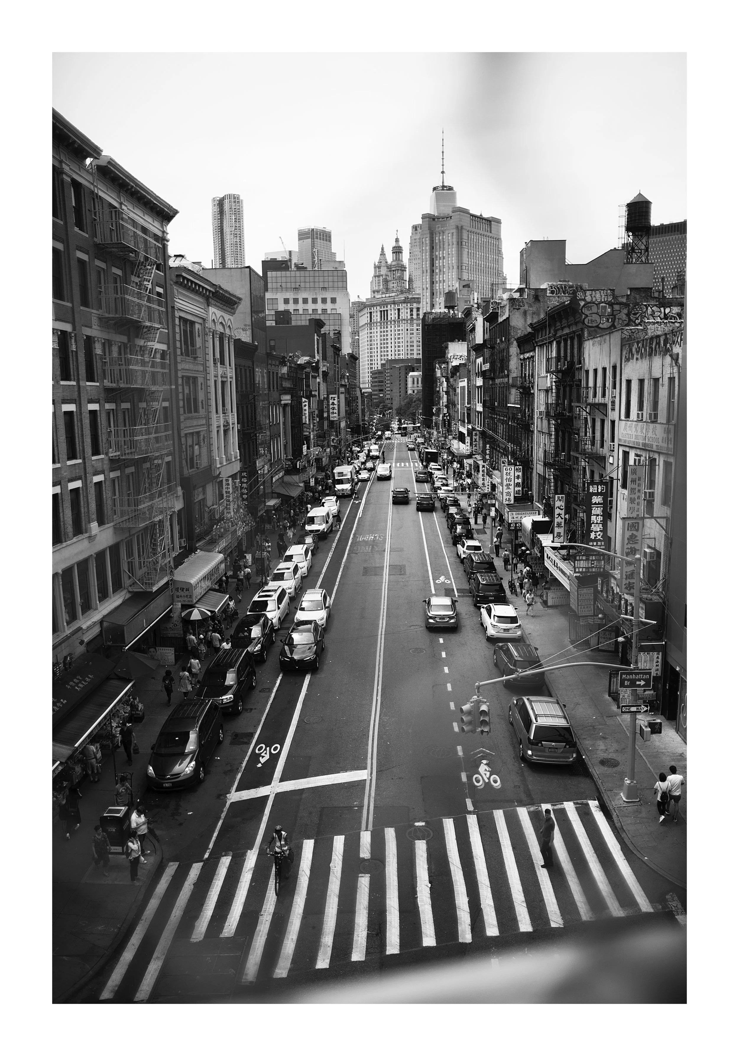 Black and white photo of a busy city street with cars, bicycles, and pedestrians, with skyscrapers in the background and a crosswalk in the foreground.