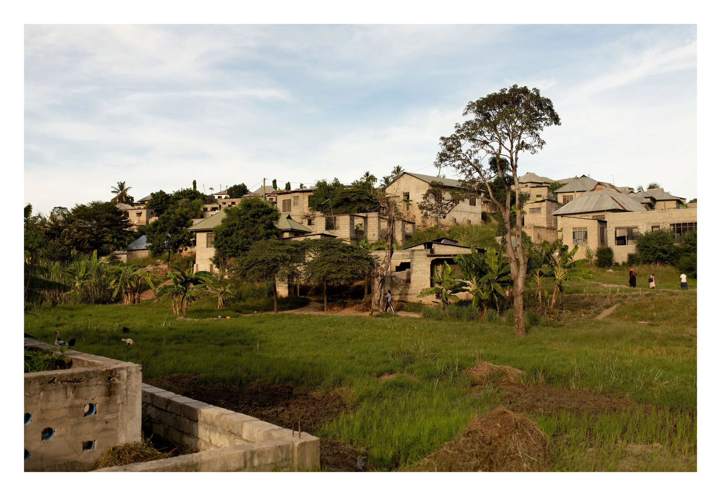 A small town or village with houses on a hillside, green trees, and a grassy area in the foreground. The houses are made of concrete blocks and have metal roofs. Several people are walking in the open grassy space, and the sky is partly cloudy.