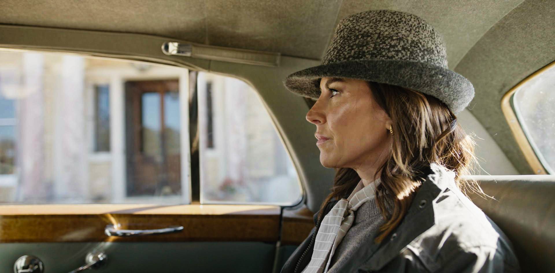 A woman with brown hair sitting in a car, wearing a gray hat and a light-colored jacket, looking out the window.