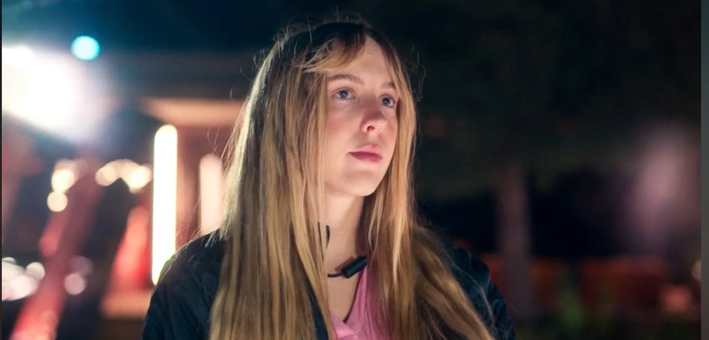 A young woman with long blonde hair and blue eyes, standing outdoors at night with blurred city lights in the background.
