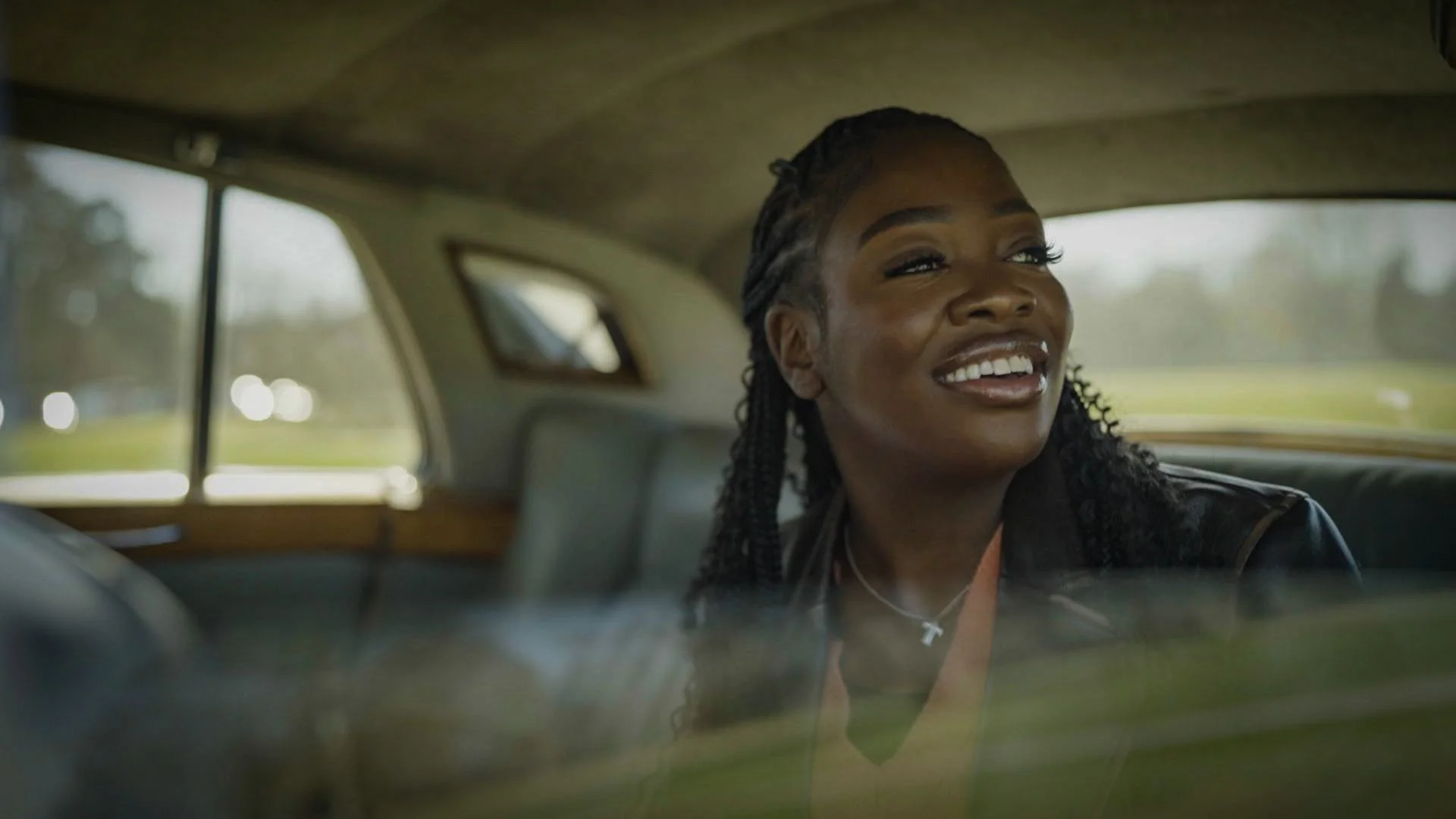 A woman with braided hair sitting in the back seat of a car, smiling and looking outside the window.