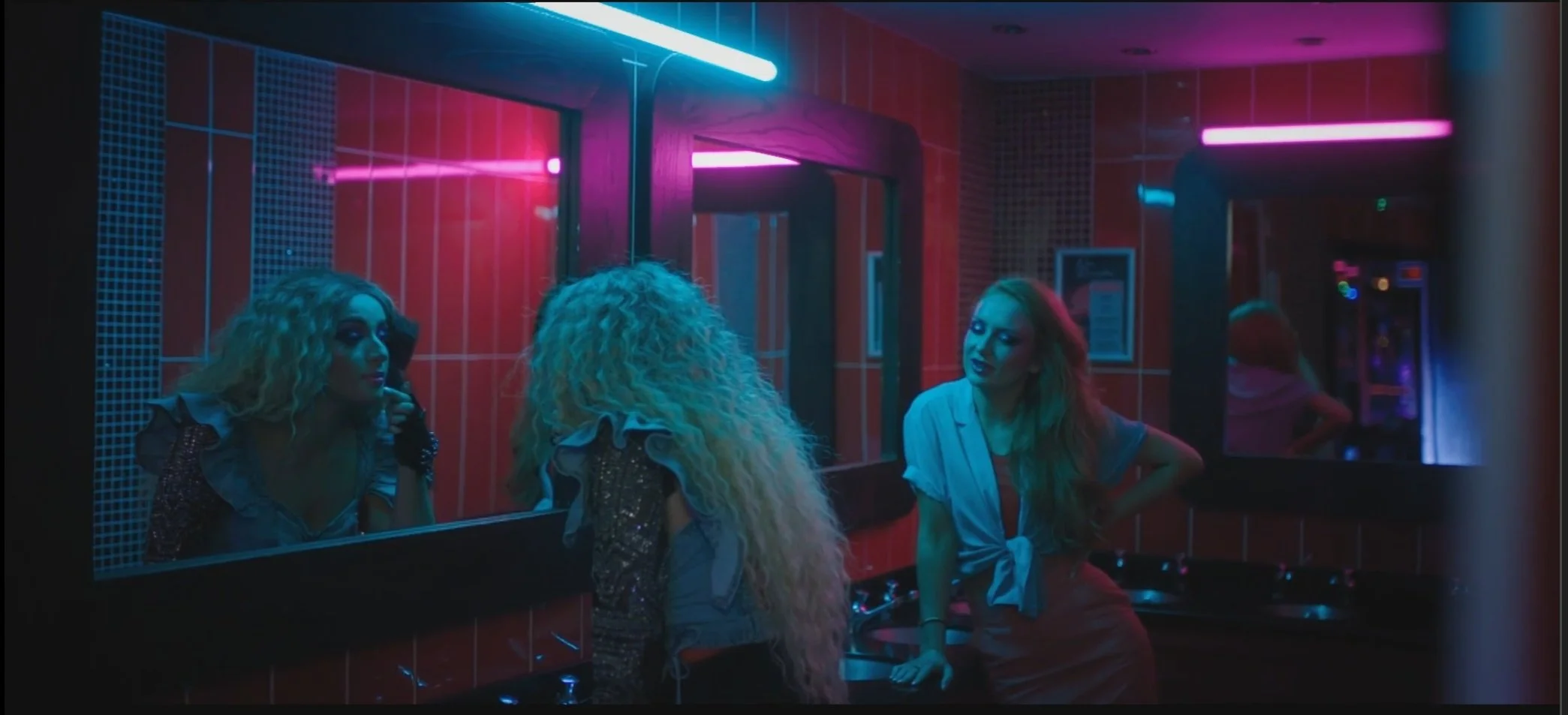 Two women with blonde curly hair and makeup are in a dimly lit bathroom with neon pink and blue lights, looking into a mirror. One is dressed in a sequined jacket, and the other in a casual shirt tied at the waist. They are engaging in conversation.