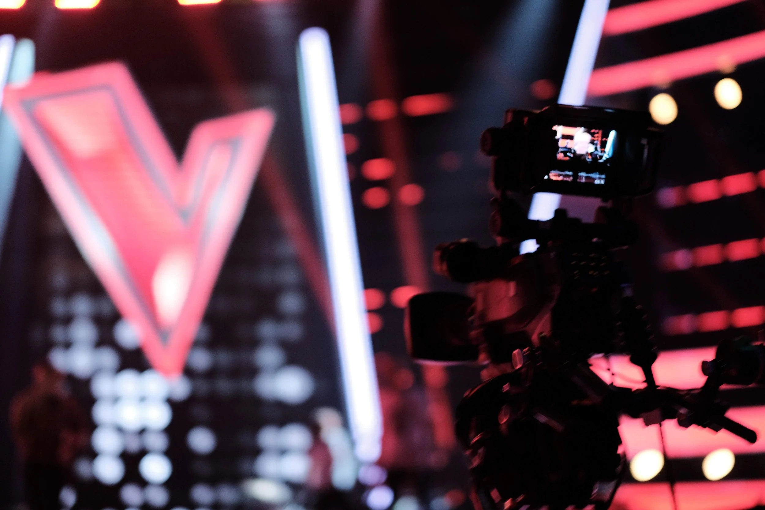 A camera mounted on a tripod recording a scene with a large V-shaped logo in the background at an event or performance.