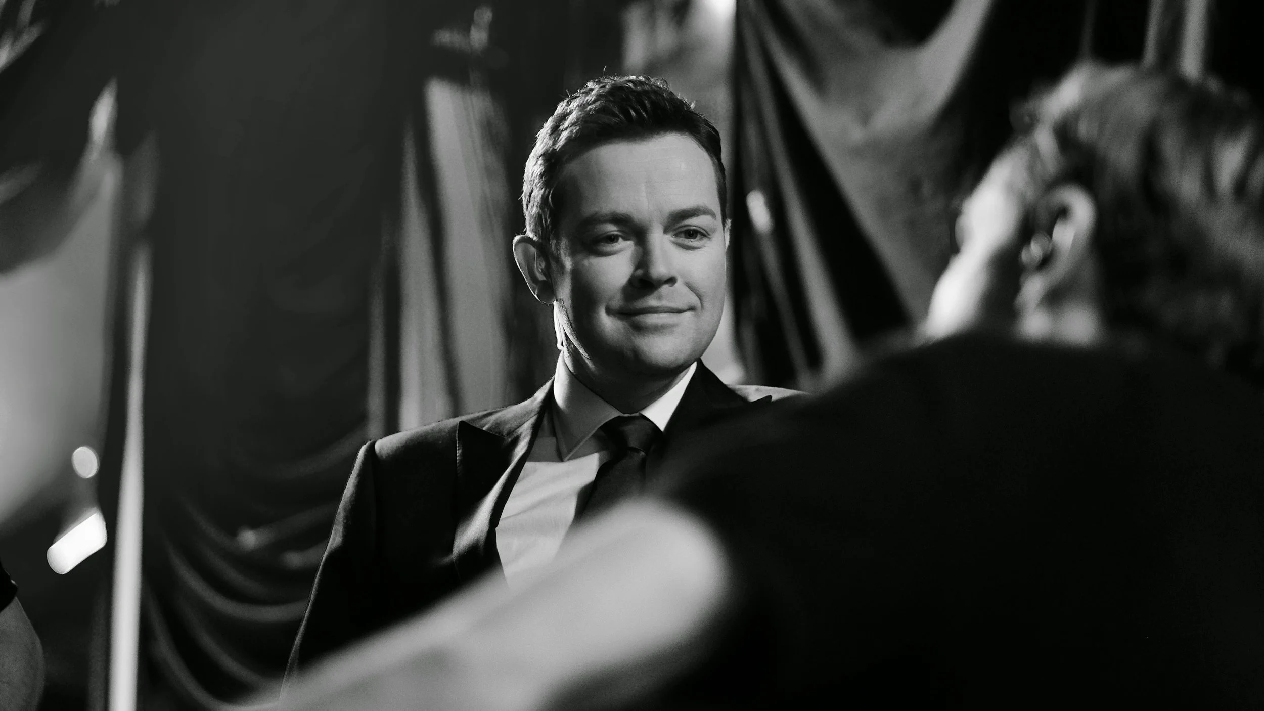 Black and white photo of a smiling man in a suit, facing an out-of-focus woman.