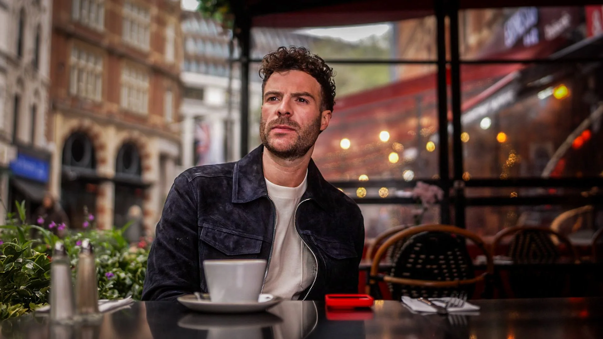 A man with dark curly hair and a beard sitting at an outdoor cafe table with a white cup, a red phone, and utensils, looking thoughtfully to his left, with city buildings and lights blurred in the background.