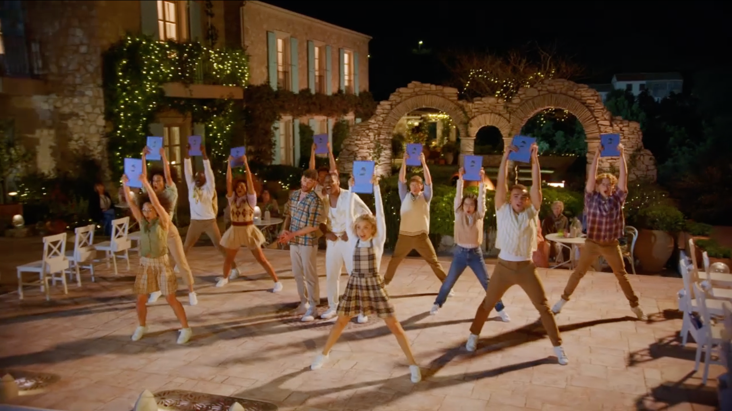 Group of children and teenagers dancing outdoors at night, holding blue certificates or papers above their heads, with festive string lights and stone archways in the background.