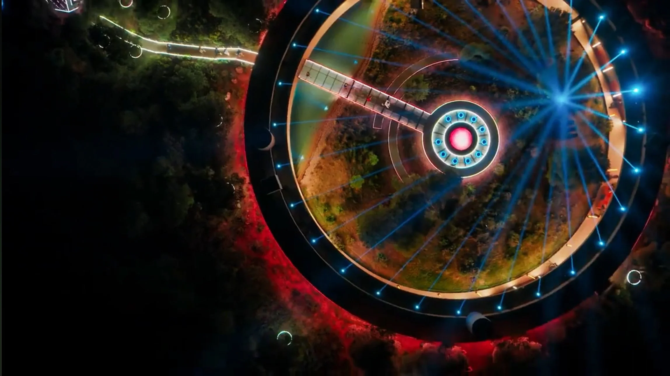 Night aerial view of a circular amusement park with colorful lights and rides, surrounded by trees and pathways.