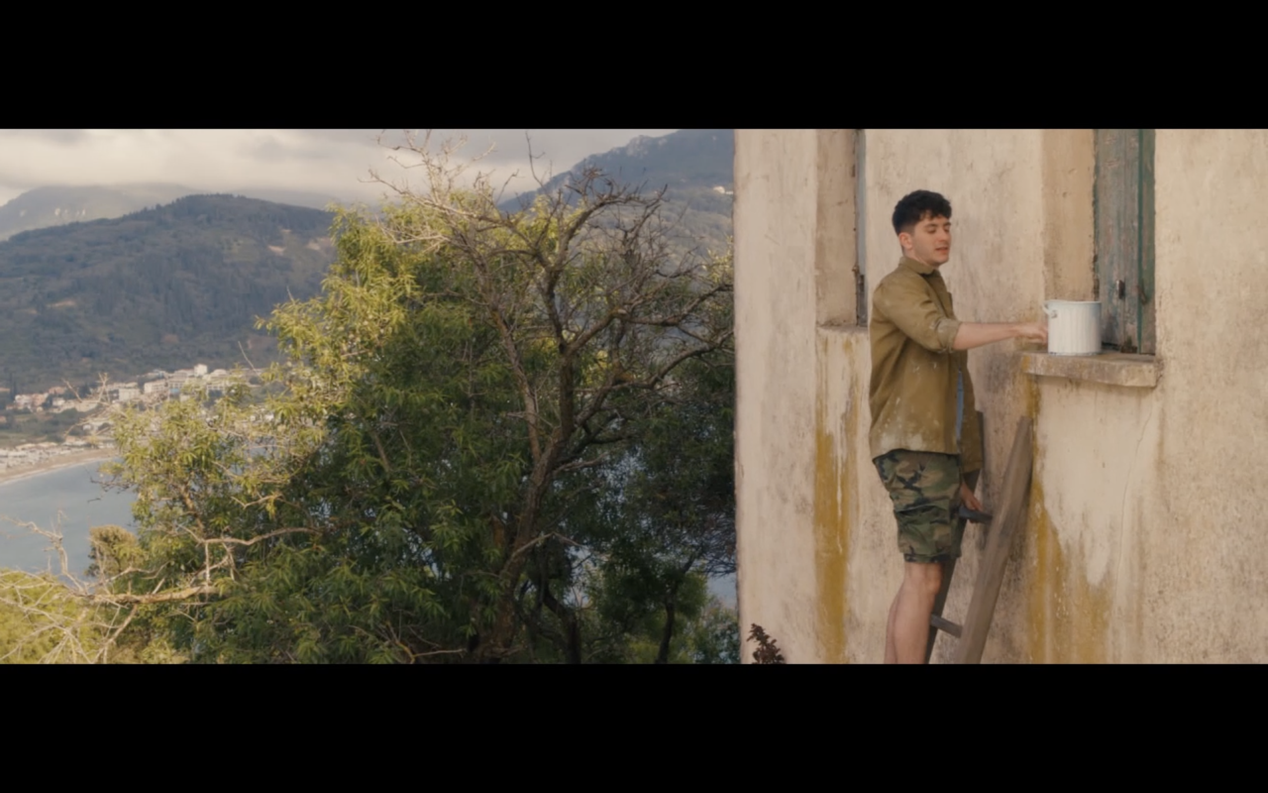 Young man standing on a ladder outside a weathered building, reaching through a window with a white bucket, overlooking a landscape with trees, water, and mountains.