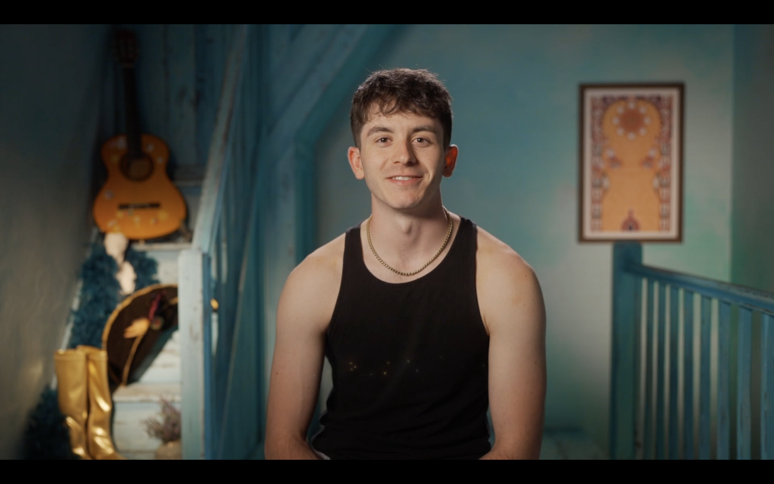 Young man with short brown hair, wearing a black tank top and gold chain, smiling in a colorful room with teal walls, a guitar hanging on the wall, and framed artwork in the background.