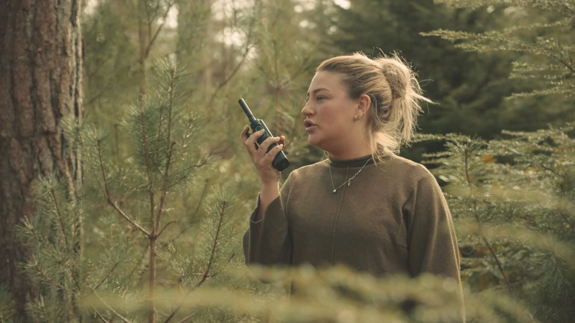 A woman standing in a forest, holding a walkie-talkie near her mouth, speaking into it, with trees and greenery in the background.