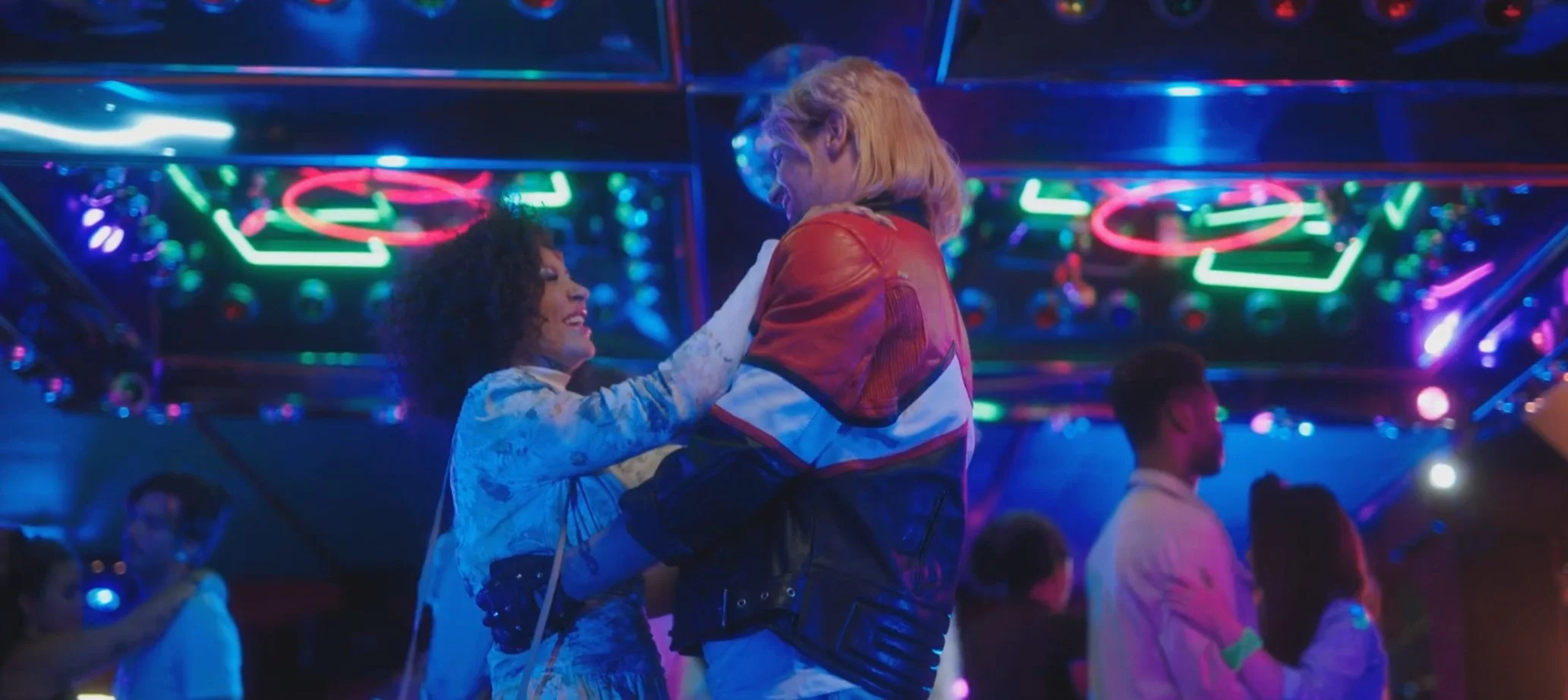 A couple dancing closely in a neon-lit club, with colorful neon signs and other people dancing in the background.