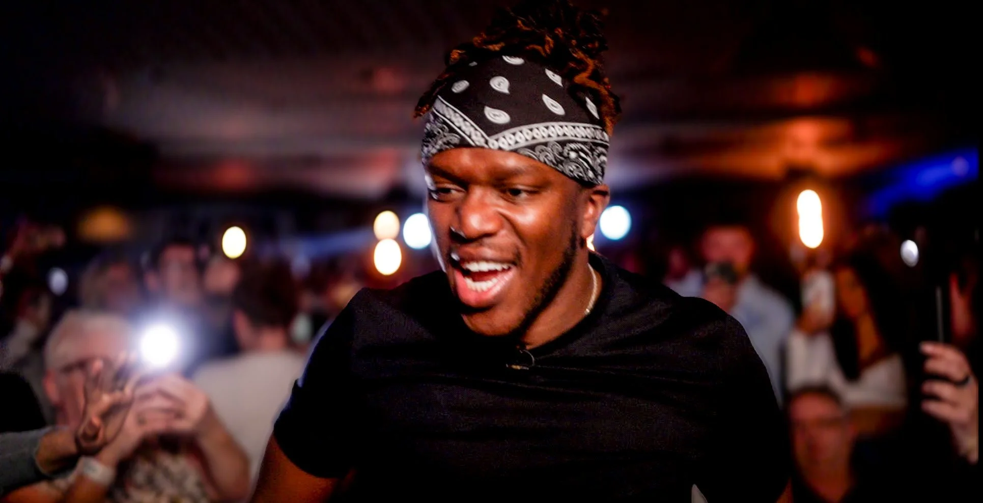 A man wearing a black bandana with a paisley pattern and a black shirt, dancing and smiling at a party or nightclub with blurred colorful lights and a crowd in the background.
