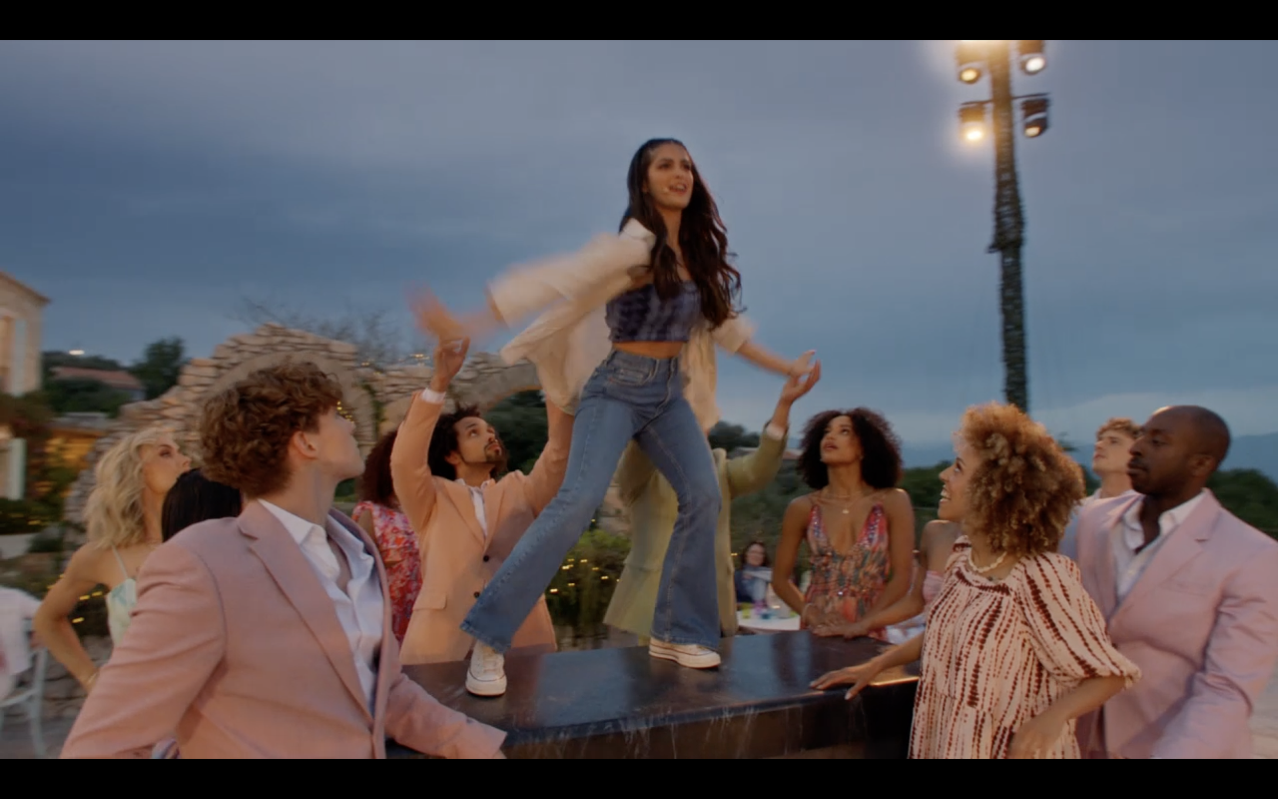 Group of diverse people around a woman standing on a table at an outdoor party during dusk, with a cloudy sky and a tall streetlight in the background