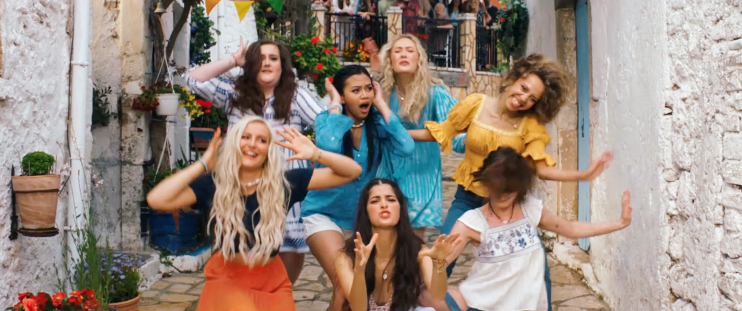 Group of young women posing playfully in a narrow outdoor alley with white stone walls and colorful flowers.
