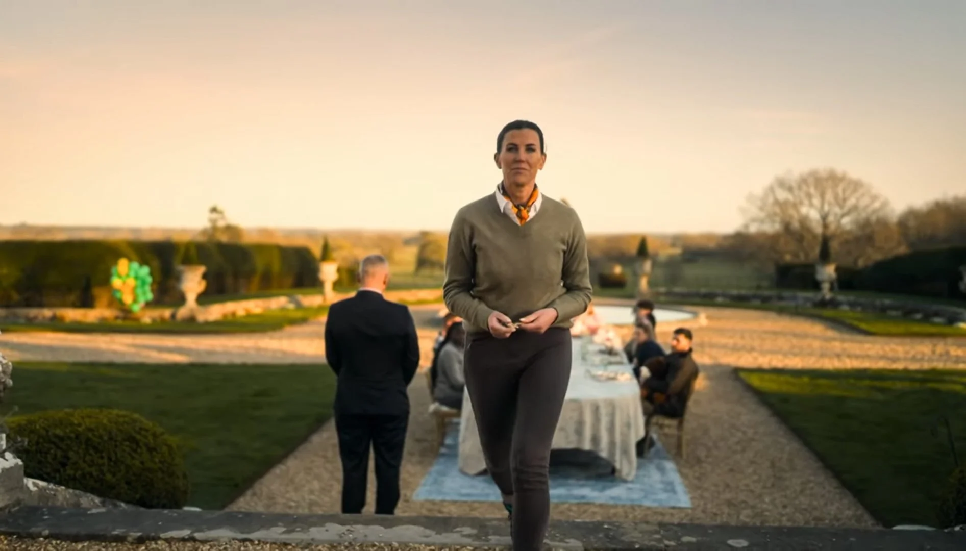 A woman walking towards the camera outdoors during a sunset, with a long dining table and several seated people in the background, on a manicured lawn with trees and decorative urns.